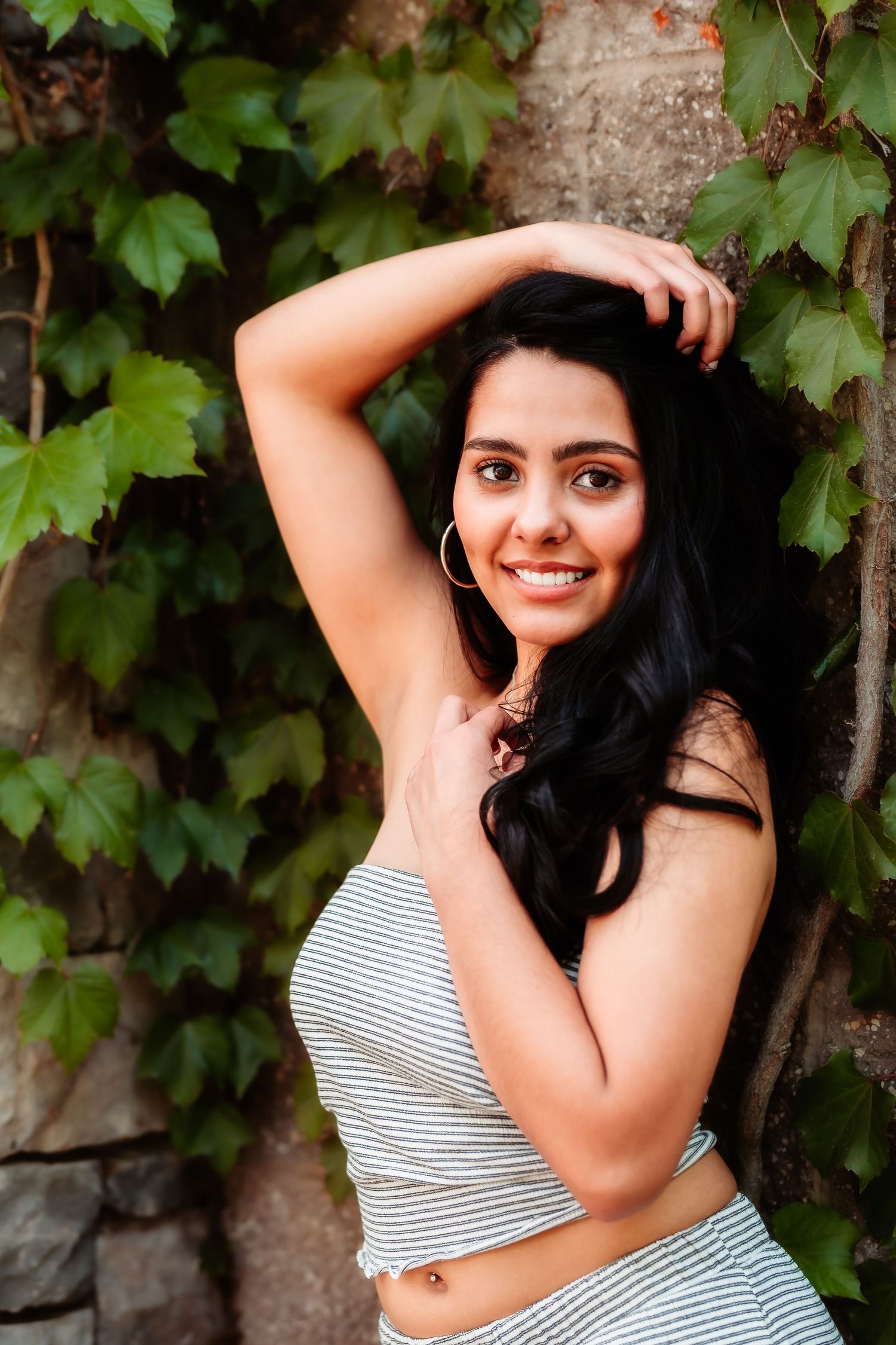 Woman in striped crop top poses against ivy-covered wall with arm raised.