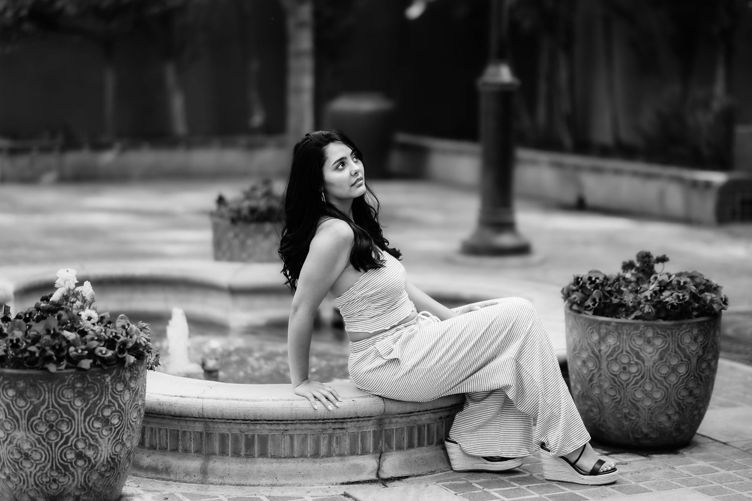 Woman in flowing dress relaxes on stone bench near decorative planters in black and white photo.