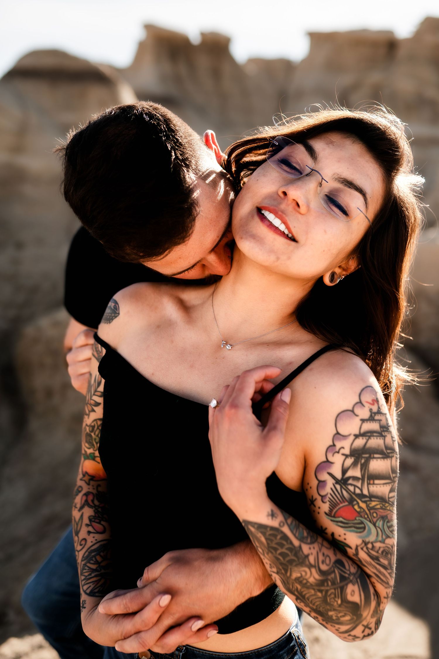 A tender romantic moment captured between two people with visible tattoos against a desert rock formation.