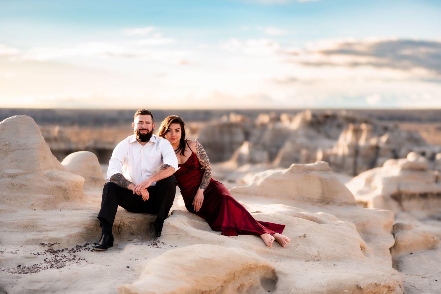 Couple in formal attire sitting on white rock formations at sunset in desert setting.