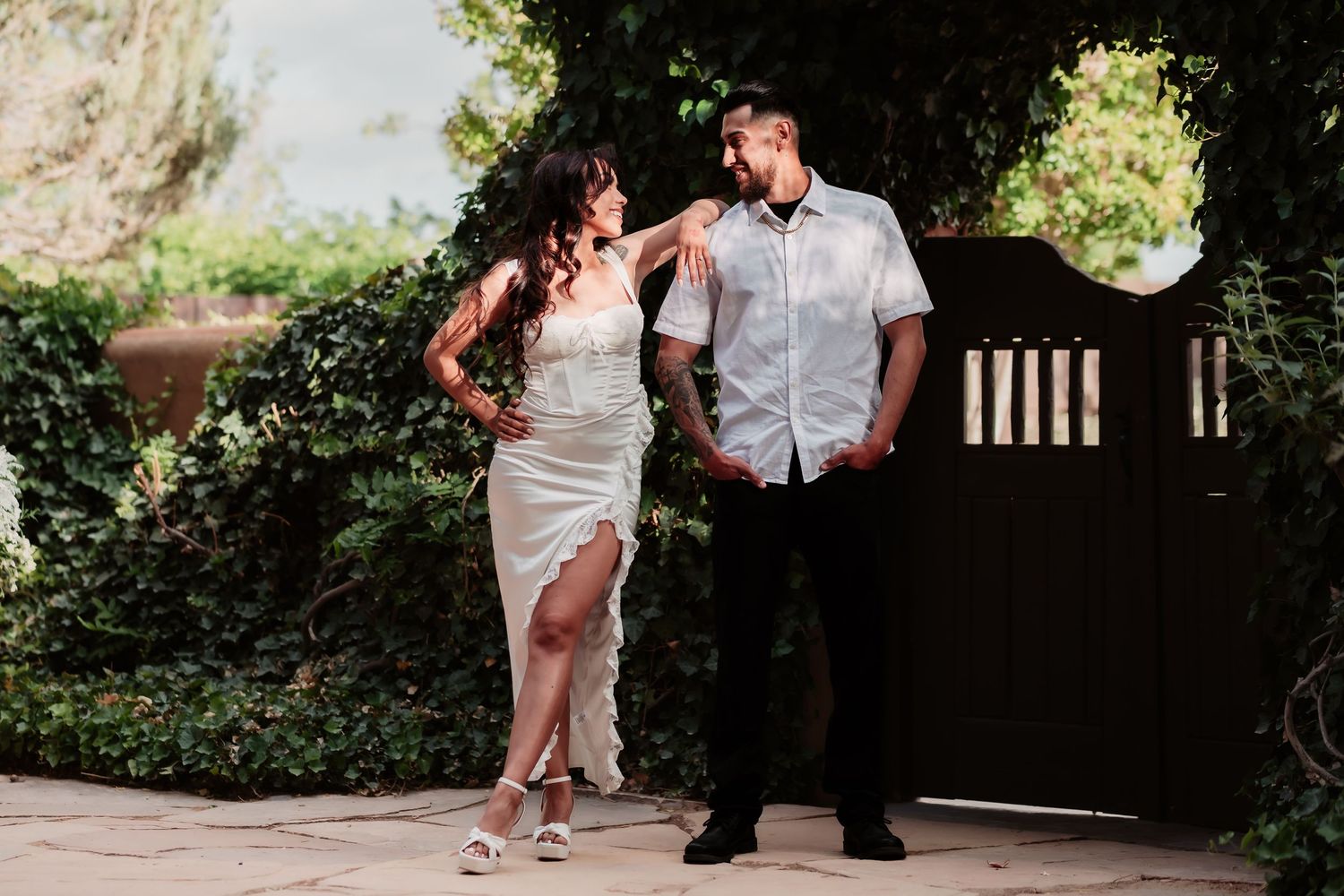 A couple in white outfits poses together by a wooden garden gate in a romantic outdoor setting.