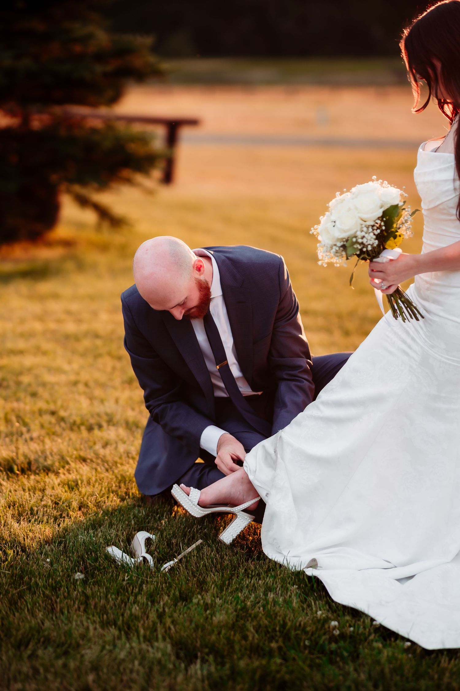 A couple in wedding attire playfully looking for something in the grass during golden hour.