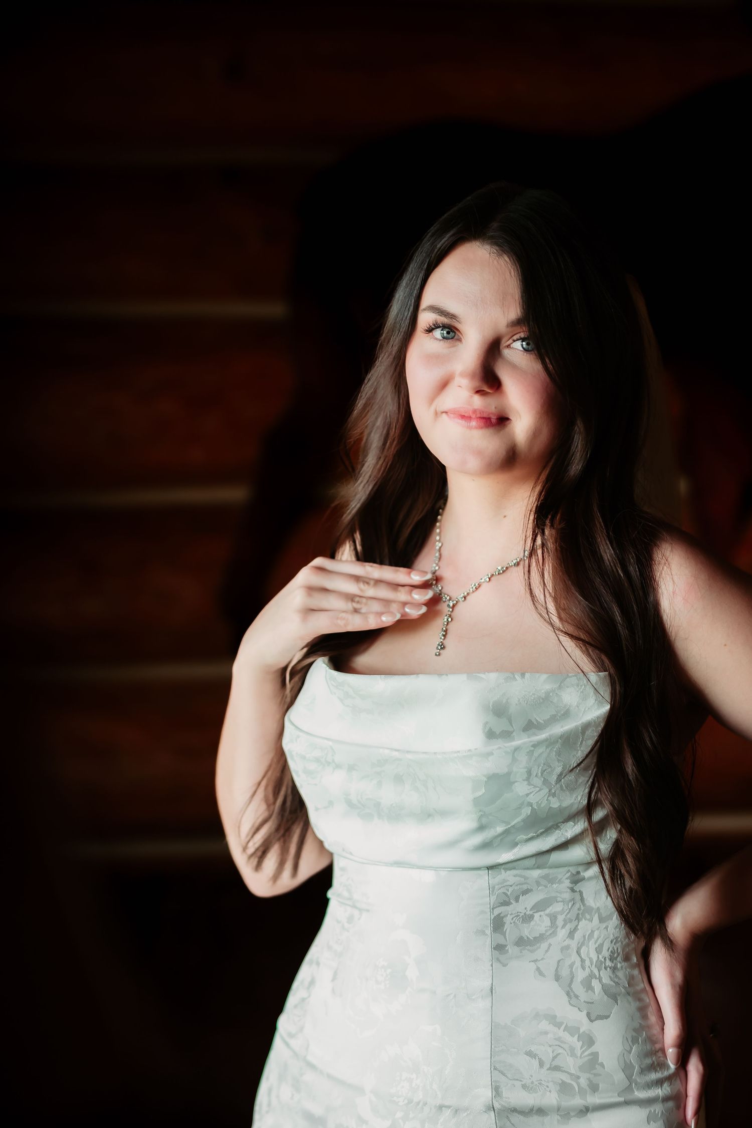 A woman in a white lace wedding dress poses against rustic wooden walls with warm lighting.