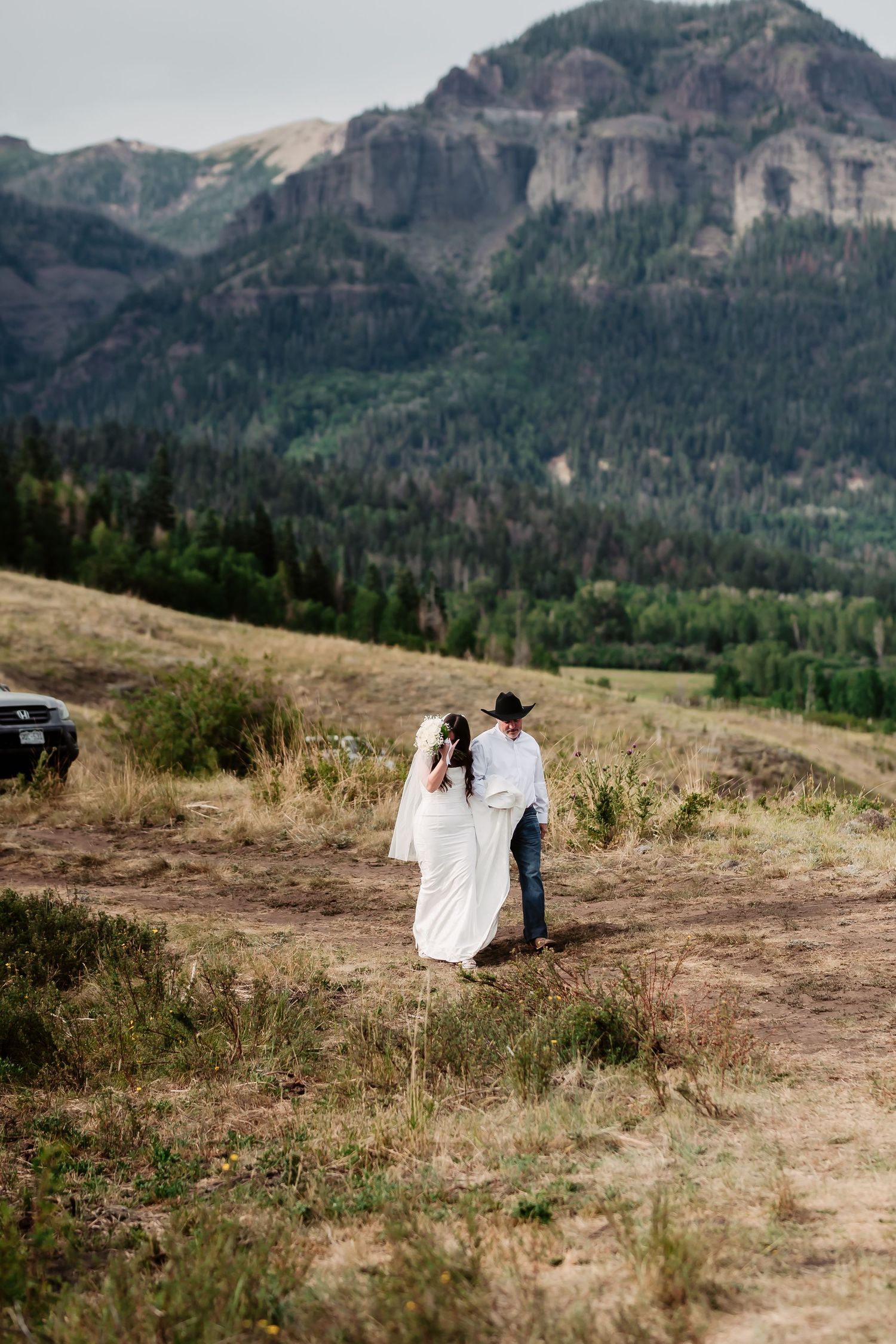 A scenic mountain wedding ceremony takes place on a grassy hillside overlooking a dramatic mountain landscape.