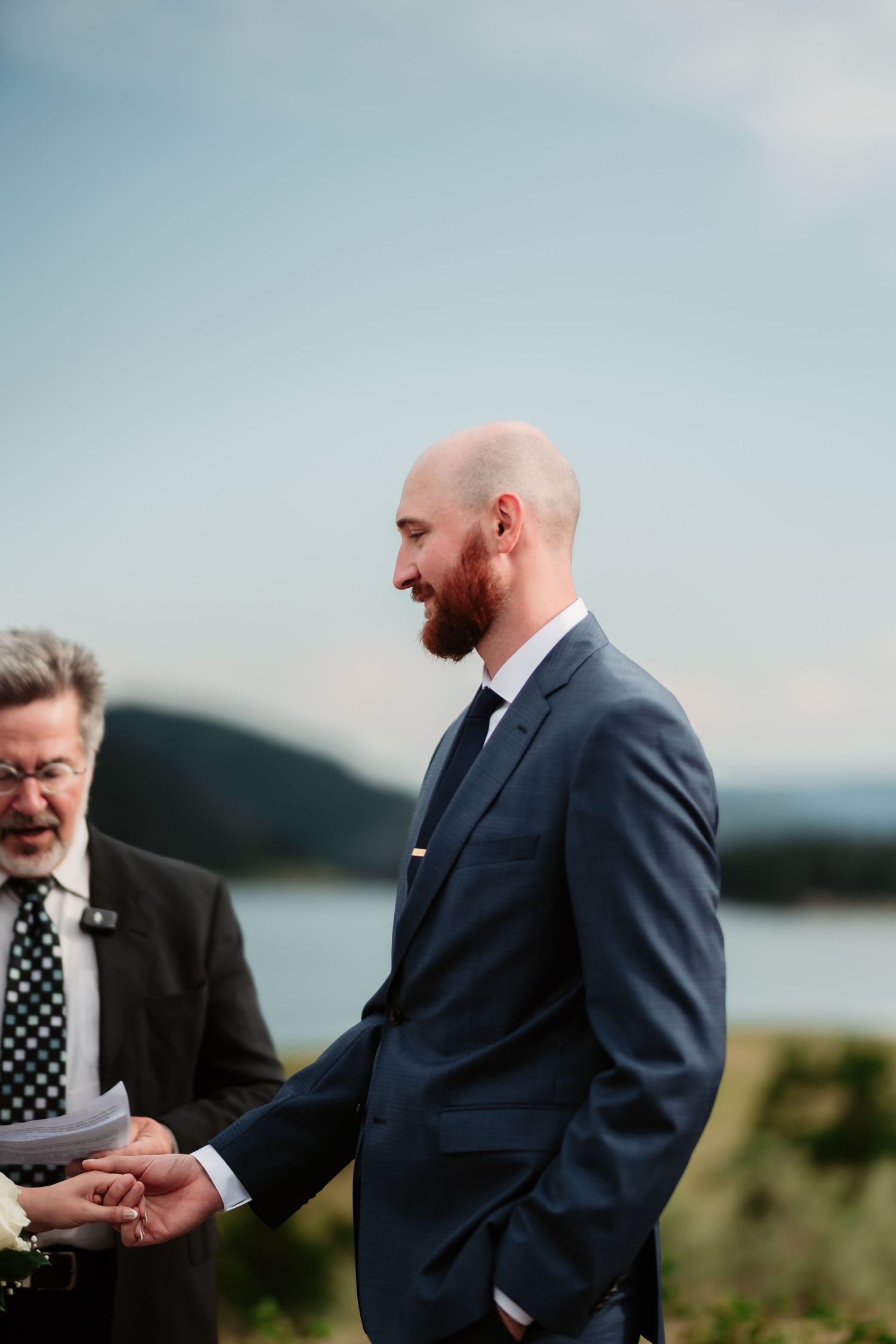 A groom in a dark suit smiles during an outdoor wedding ceremony with mountains and water in view.