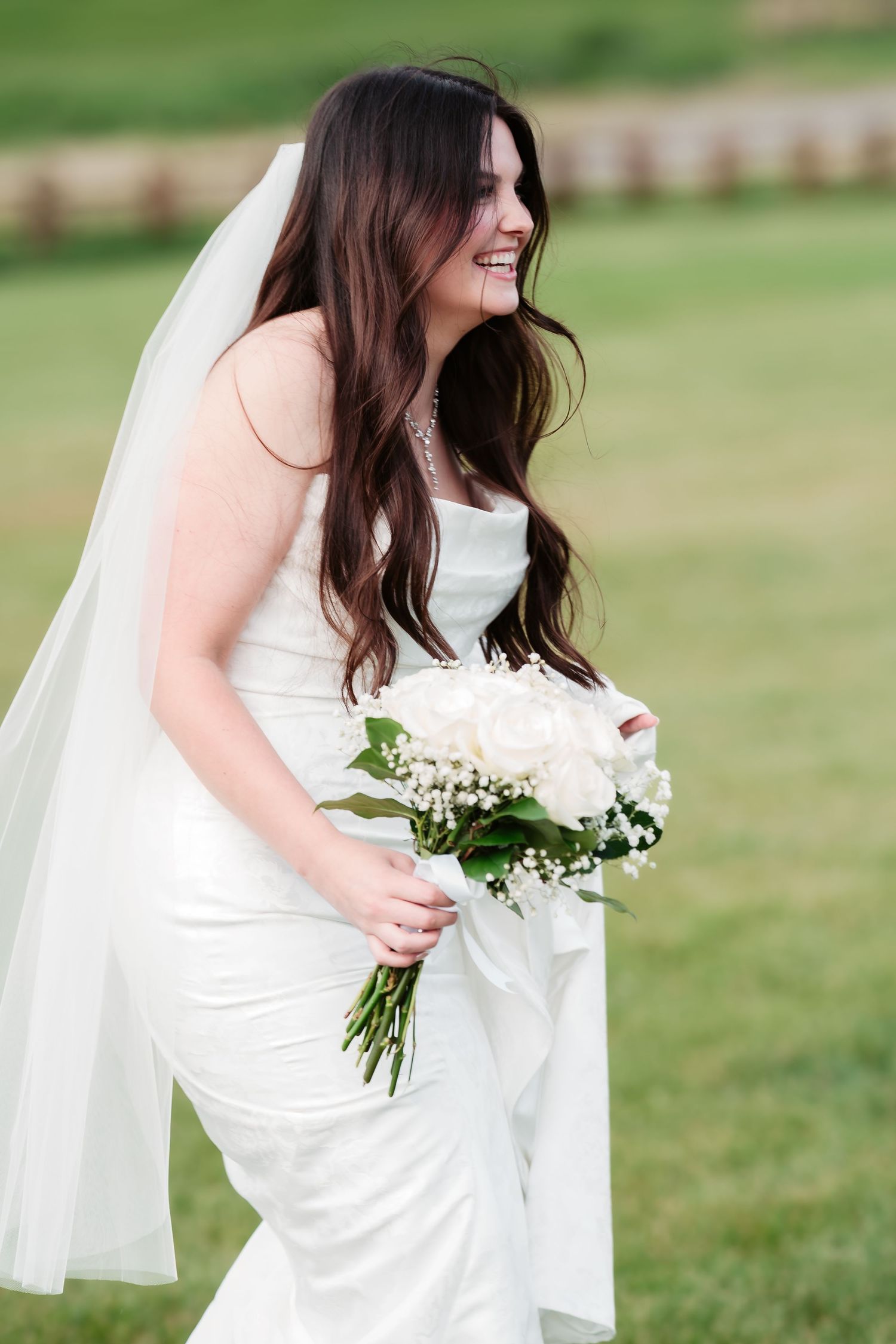 A bride in a fitted white gown and long veil holds a white floral bouquet while smiling on her wedding day.