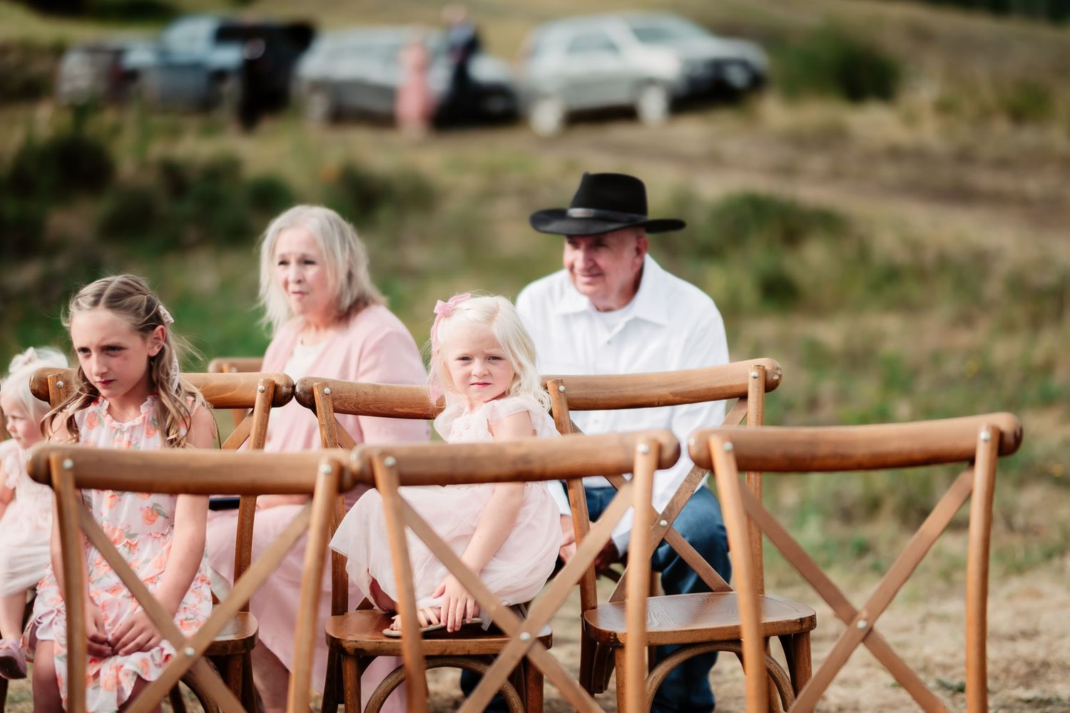 Wedding guests seated in wooden cross-back chairs at an outdoor ceremony in a rustic field setting.