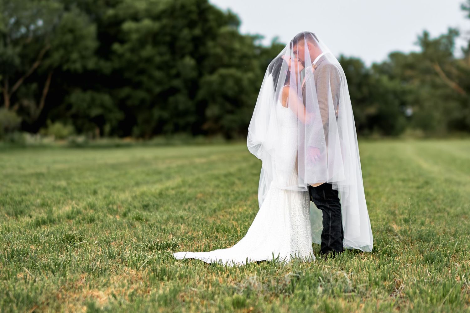 Artistic wedding photo with bride's veil flowing in the wind on a grassy hilltop.