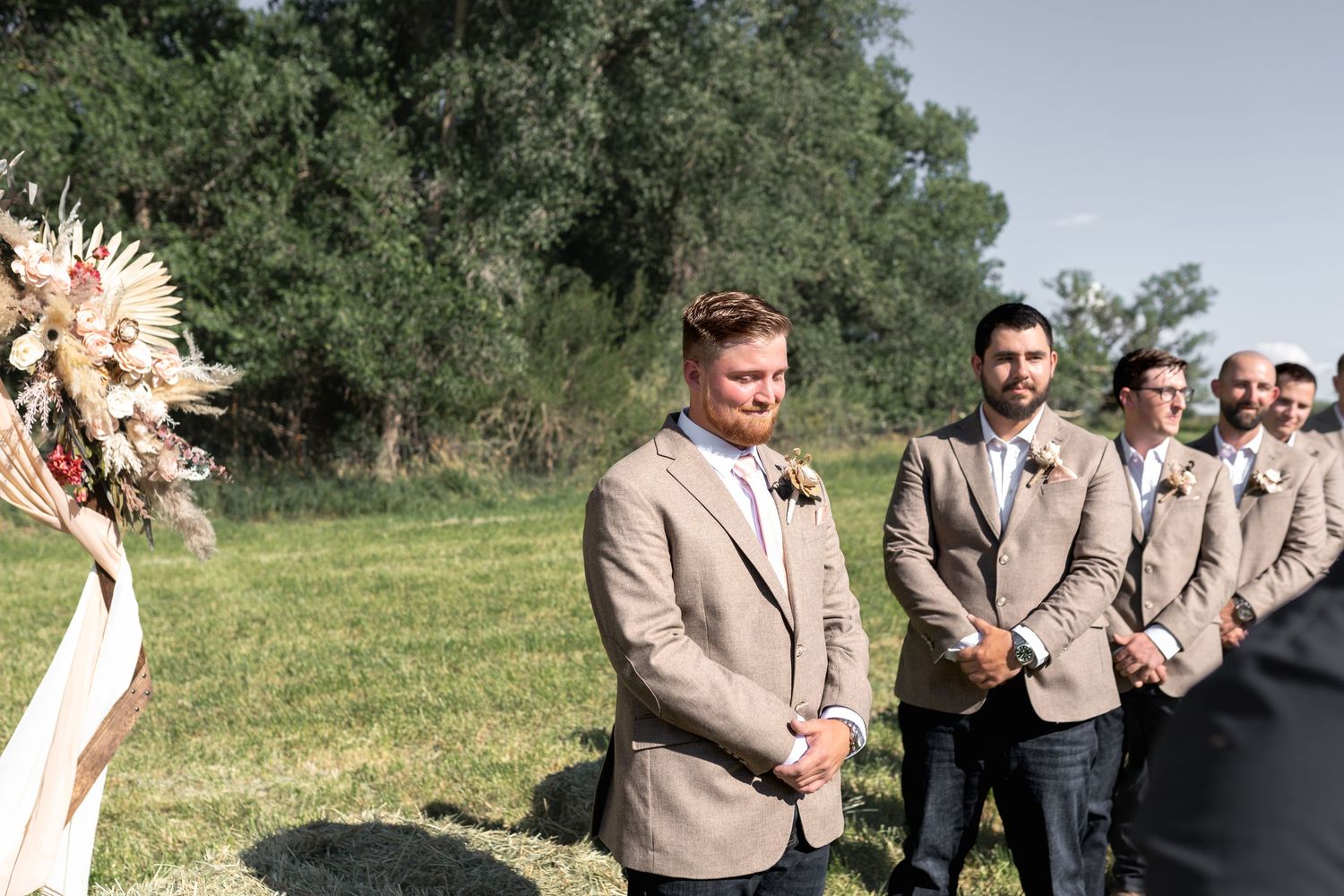 Groomsmen in tan suits stand attentively during an outdoor wedding ceremony.