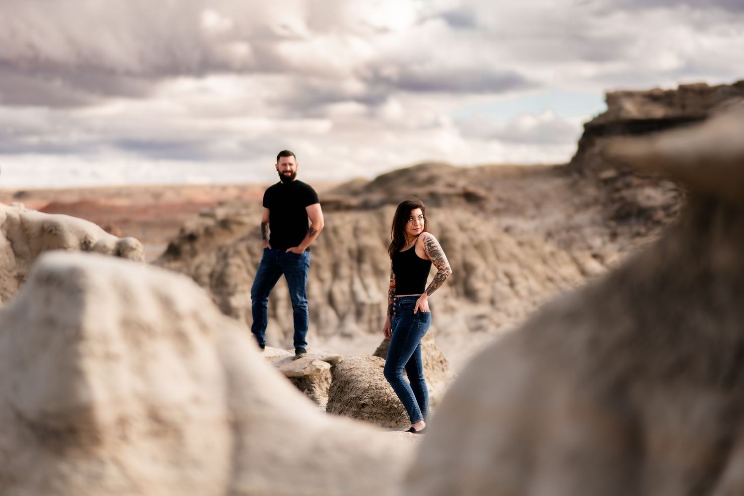 Two people standing on rugged rock formations in a desert landscape under dramatic cloudy skies.