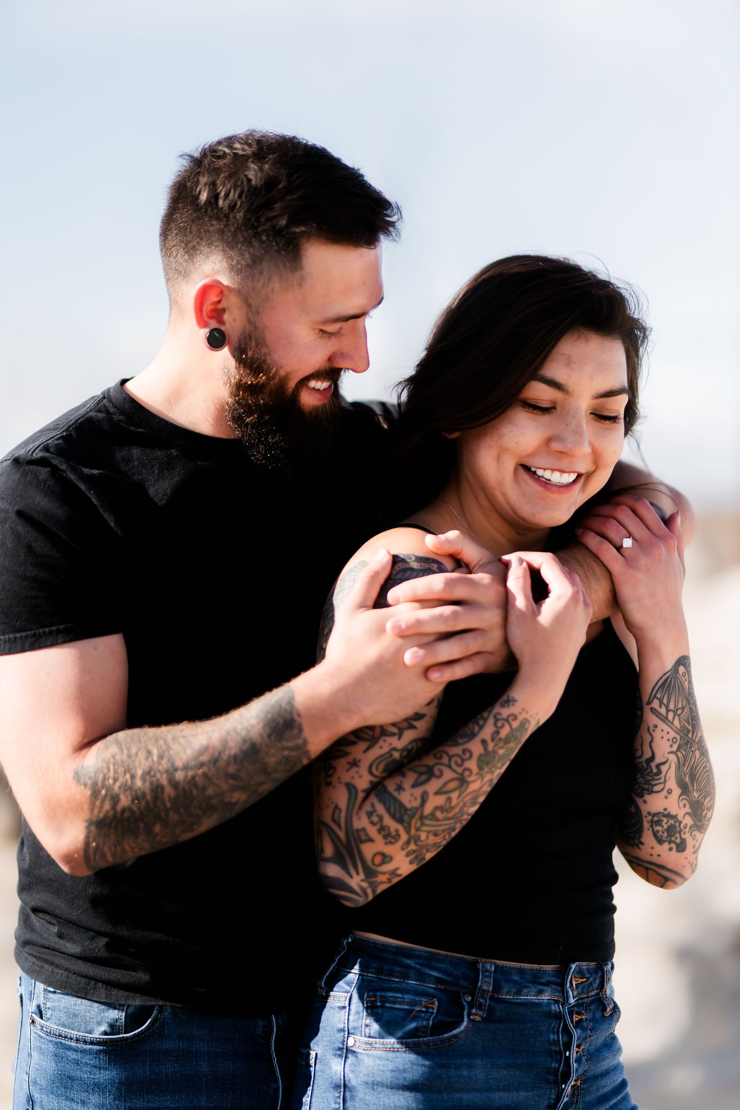 Two people embrace intimately in matching black shirts and jeans, showing off their arm tattoos.