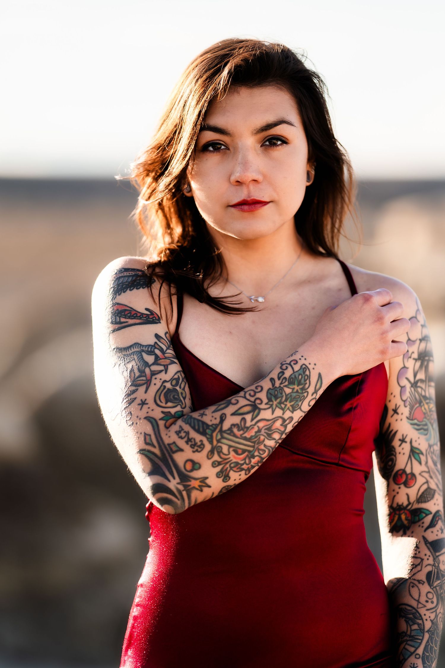 Woman in red sleeveless dress with intricate floral and decorative tattoo sleeves poses outdoors at sunset.