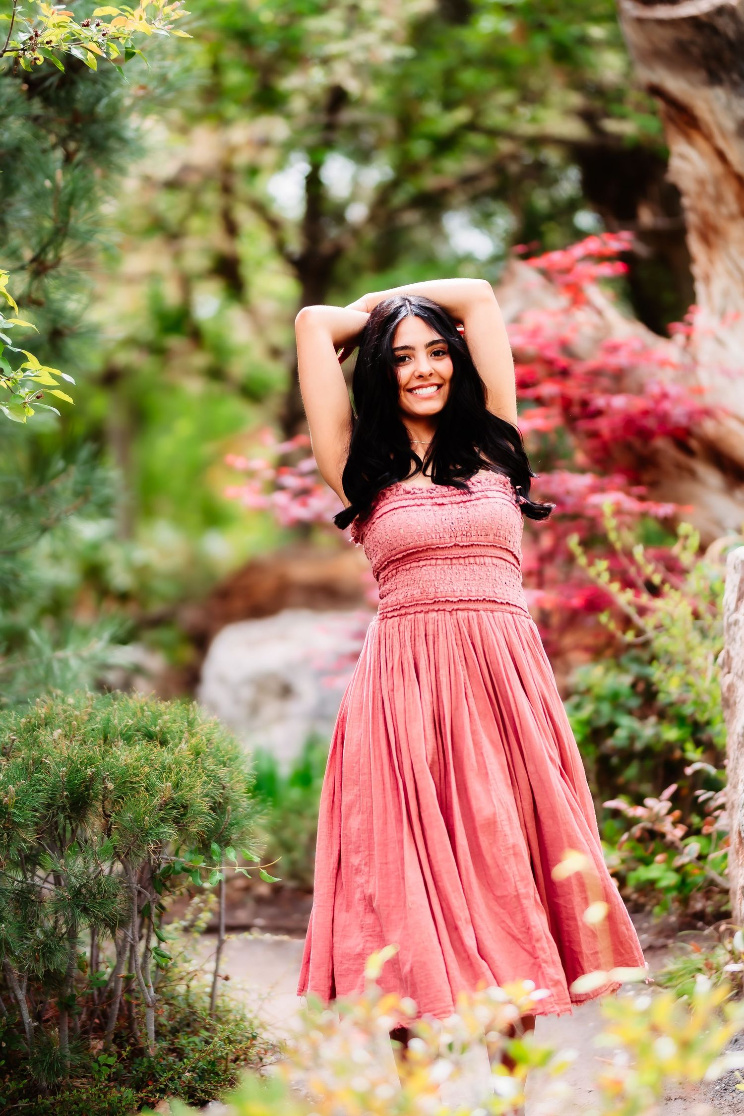 A pink flowy dress stands out against lush garden foliage with colorful red maples in the background.