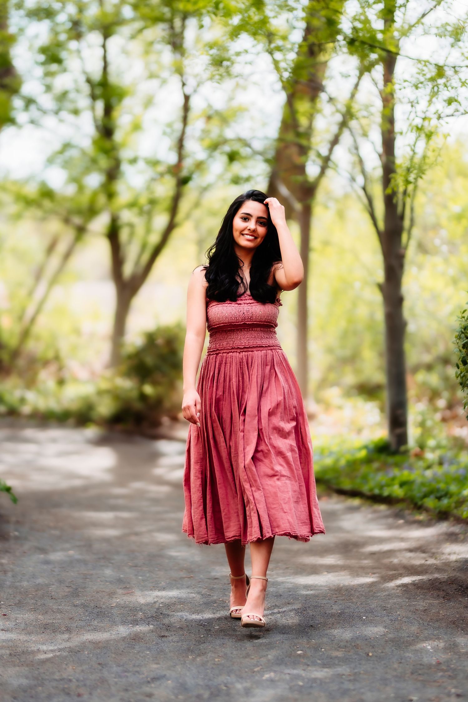 A woman in a flowing red sundress walks along a tree-lined path on a sunny day.