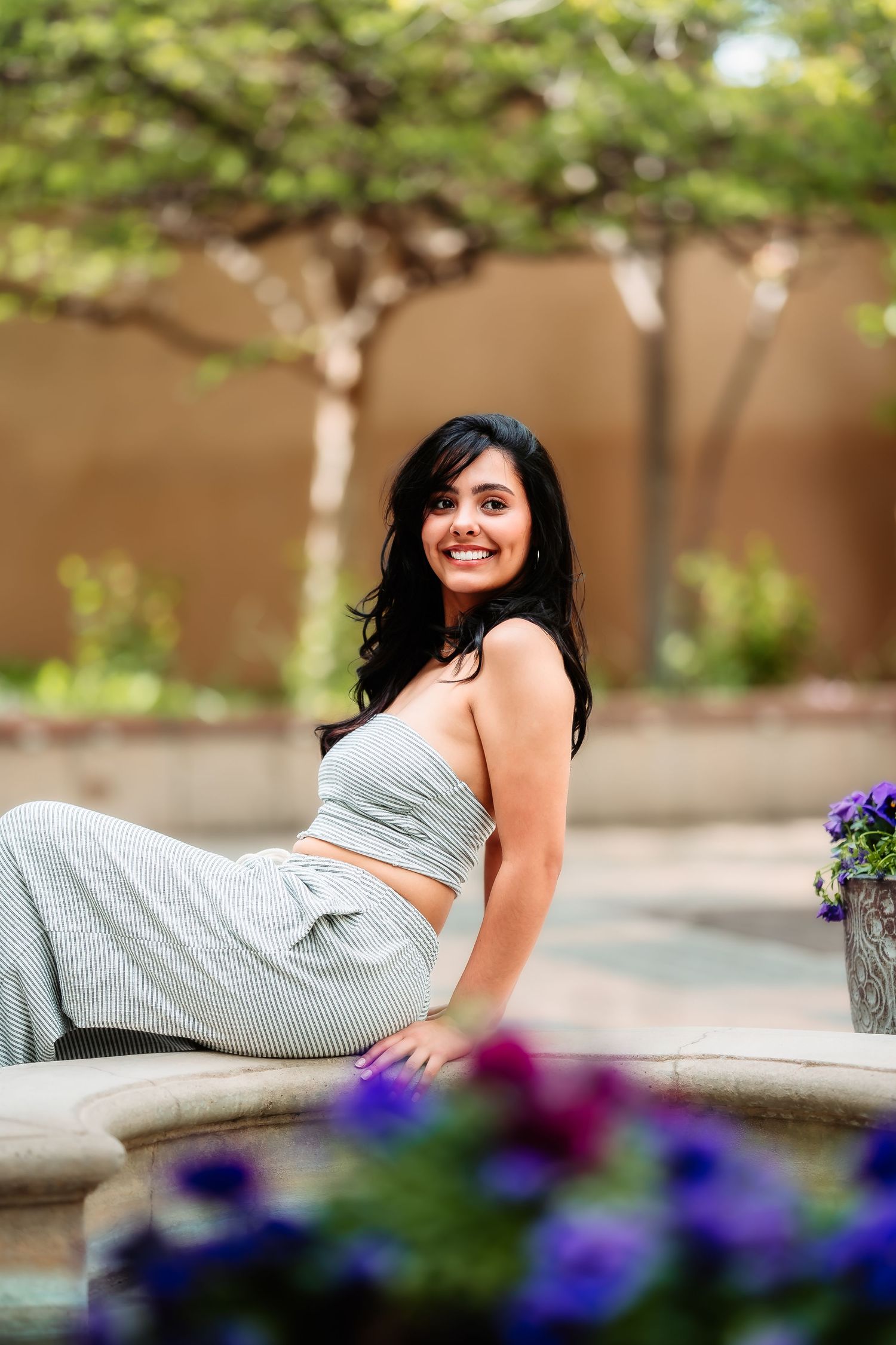 Woman in gray two-piece outfit relaxing by outdoor fountain with purple flowers in stone planters.