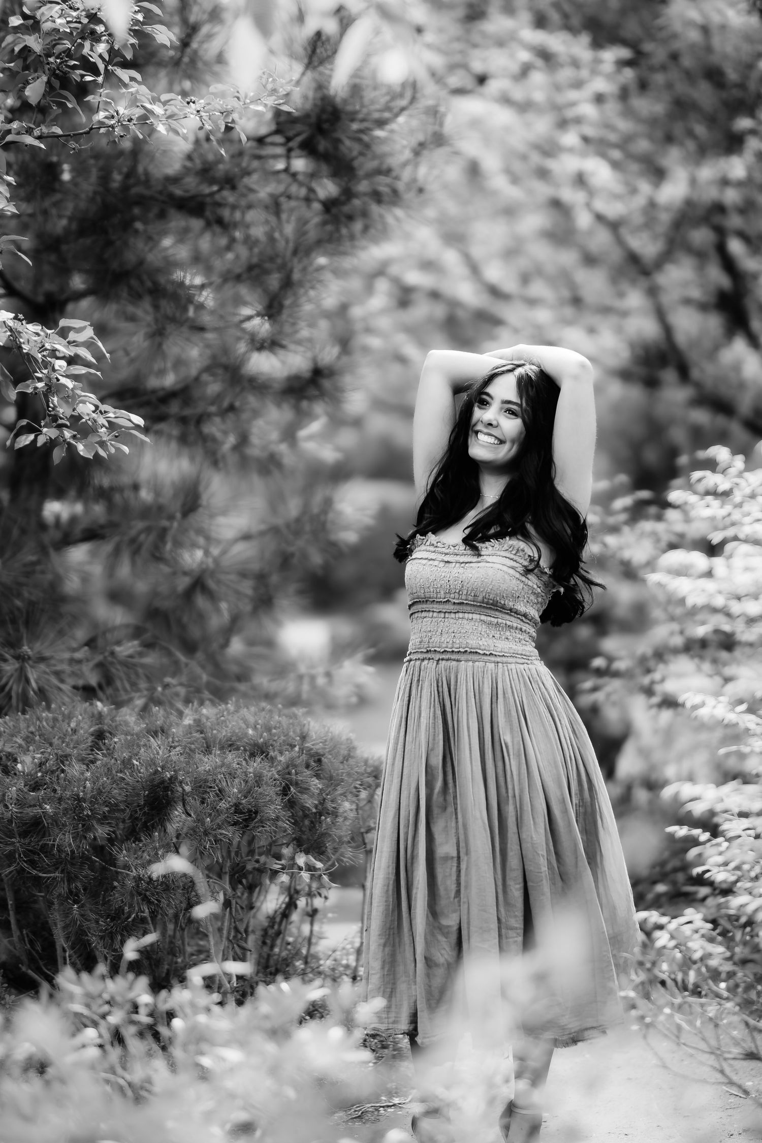 A black and white portrait of someone in a flowing dress posing gracefully in a garden setting.