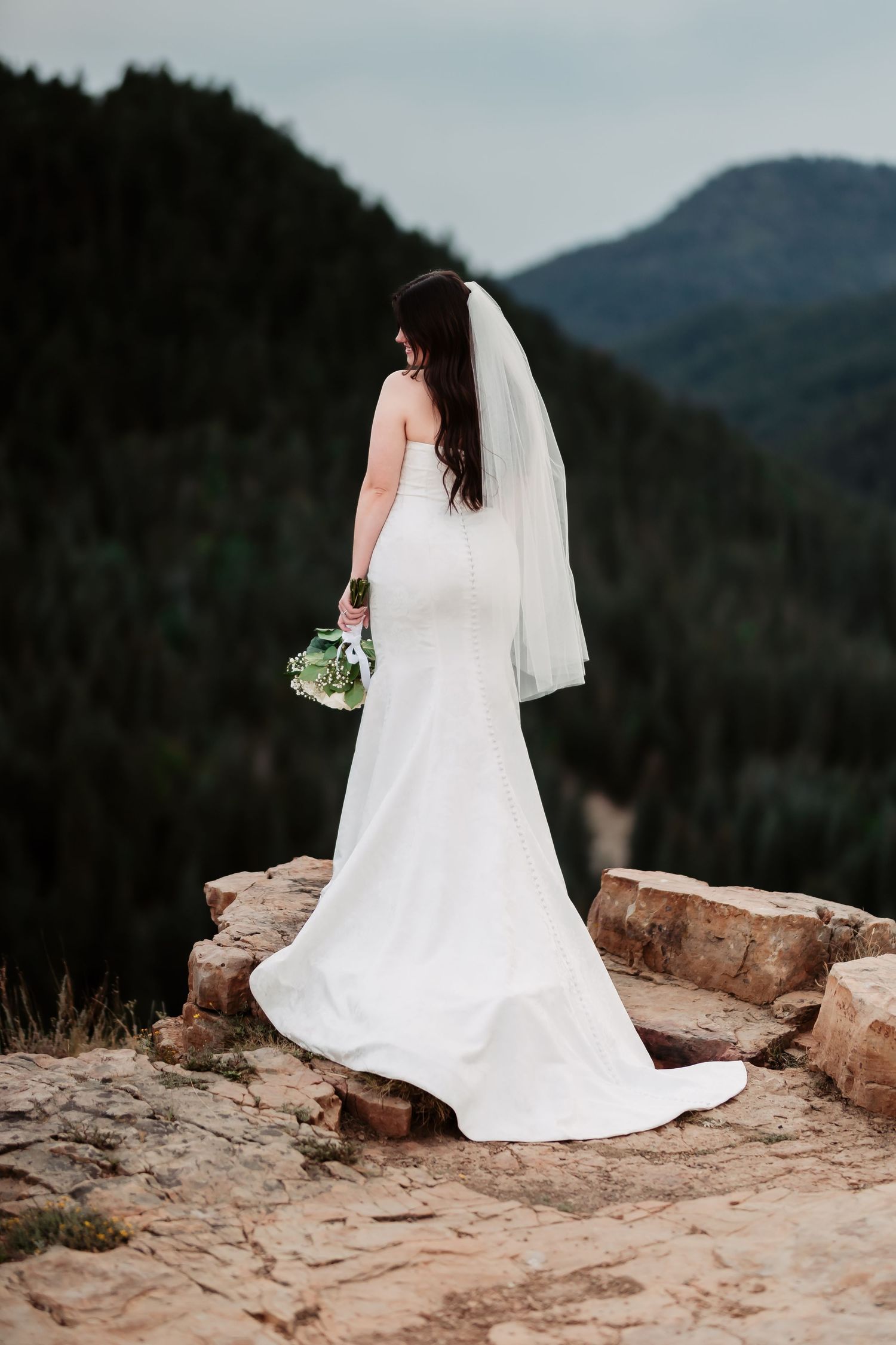 Bride in flowing white gown sits peacefully on rocky mountain ledge with scenic backdrop.
