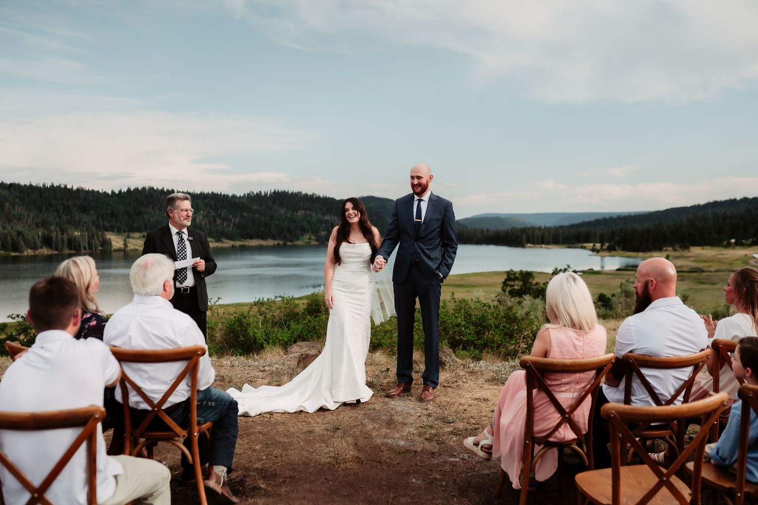 Intimate outdoor wedding ceremony overlooking a scenic lake with mountains in the background.