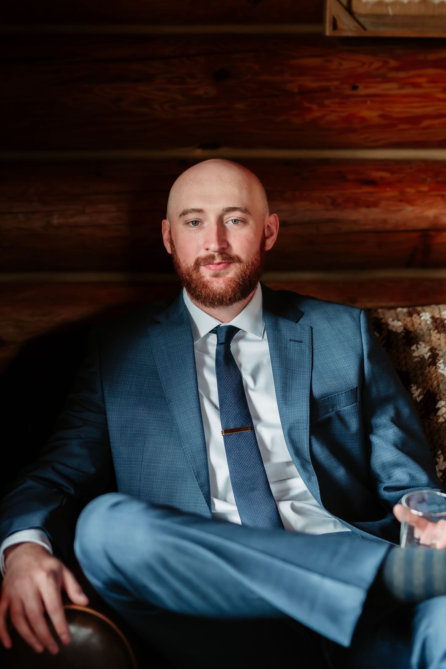 Formal portrait in blue suit seated on leather couch in rustic cabin setting.