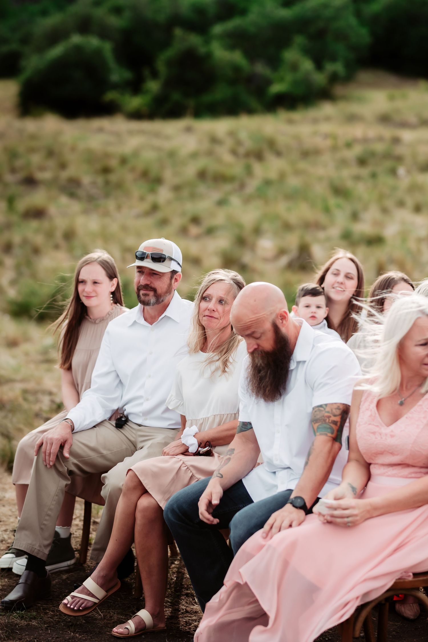 Wedding guests seated in rustic chairs outdoors smile and laugh during an emotional ceremony moment.