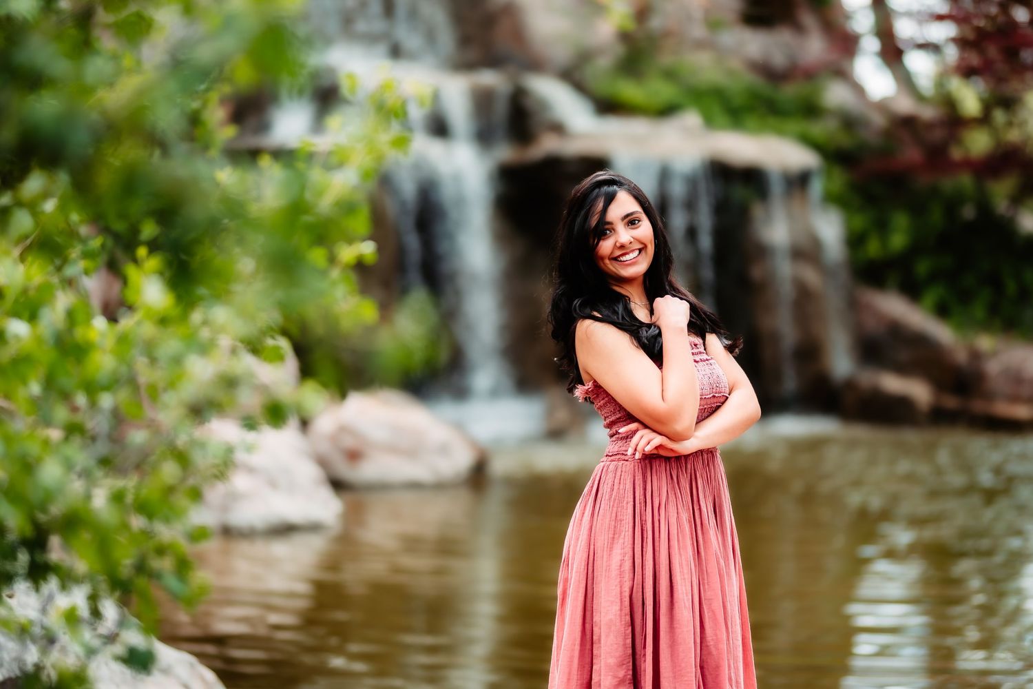 Person in coral dress poses elegantly in front of cascading waterfall and pond in lush garden setting.