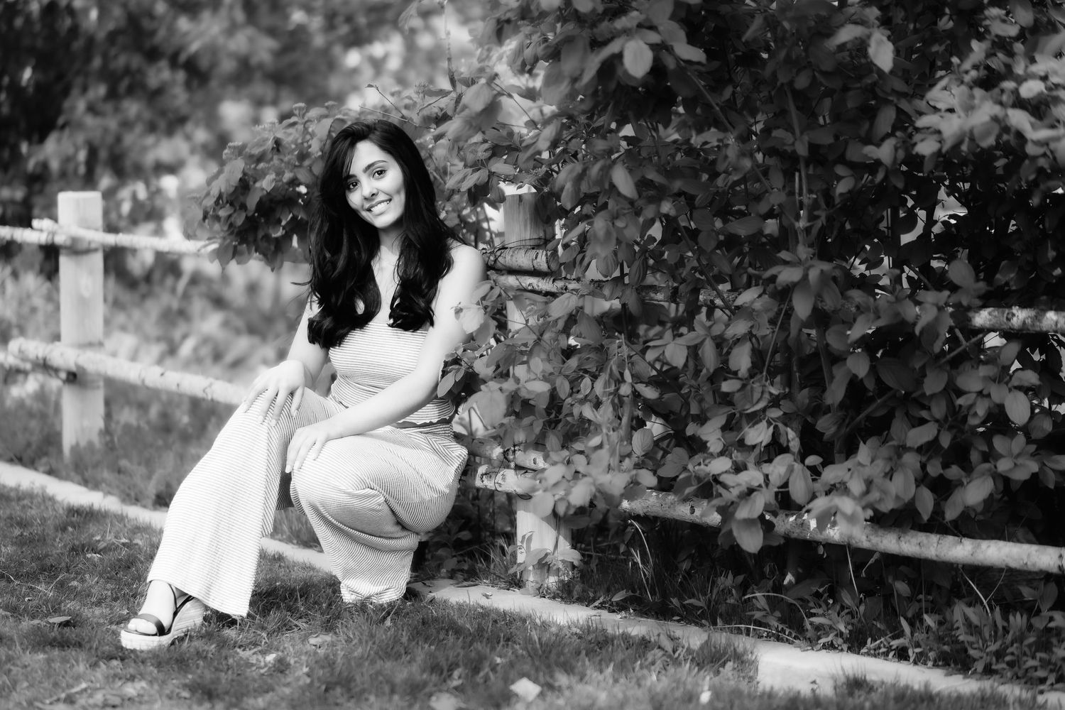 Black and white photo of woman in casual attire crouching by wooden fence outdoors.