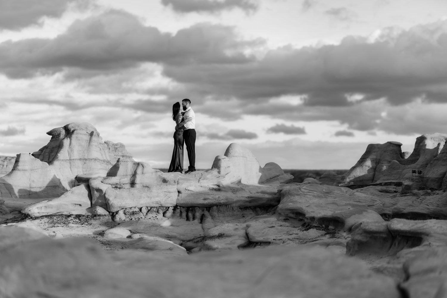 Black and white photo of couple standing on dramatic badlands rock formations under cloudy sky.