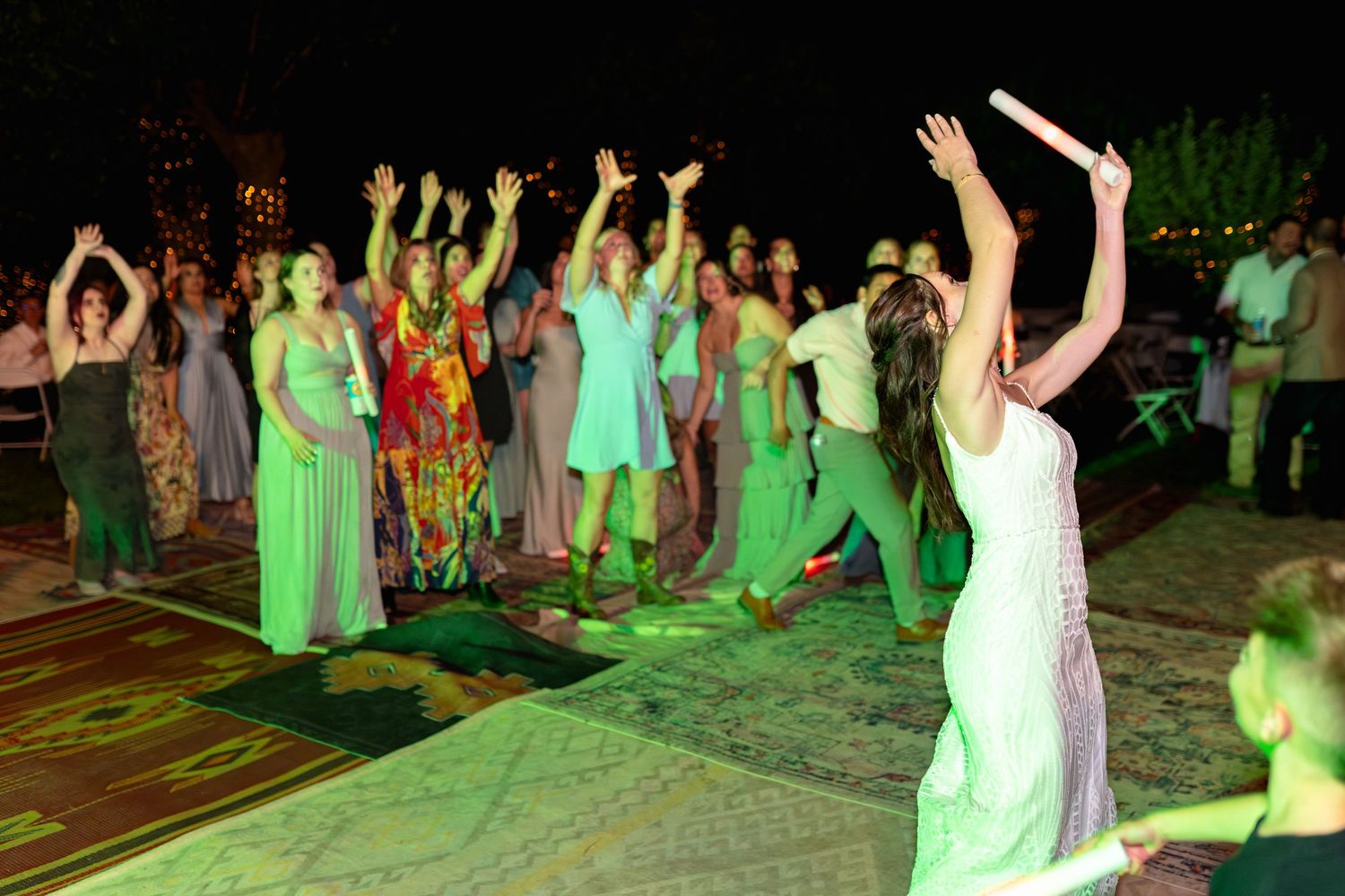Wedding guests in green dresses dance while the bride throws her arms up on an illuminated dance floor.
