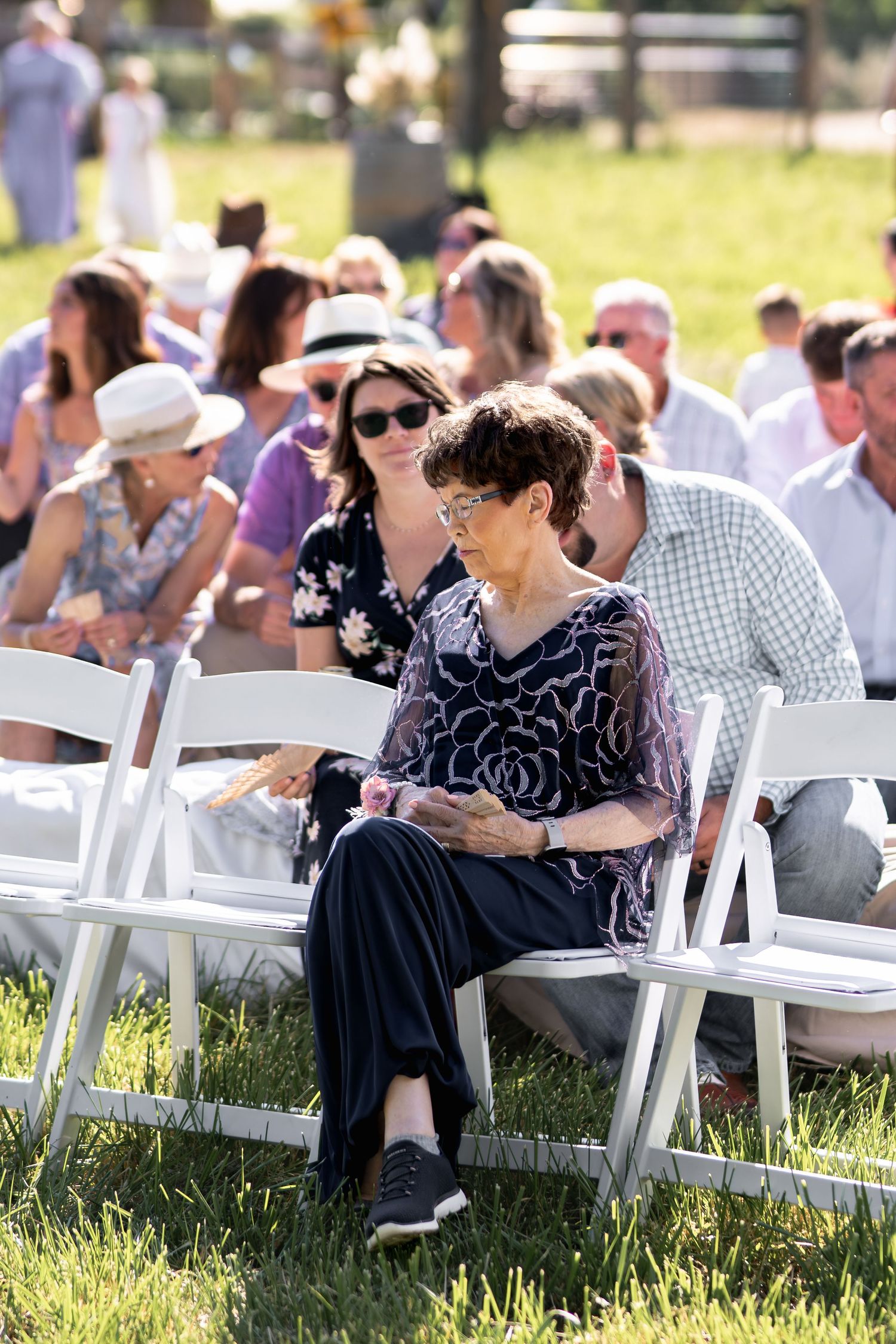 Wedding guests seated on white chairs on a grassy lawn during an outdoor ceremony.