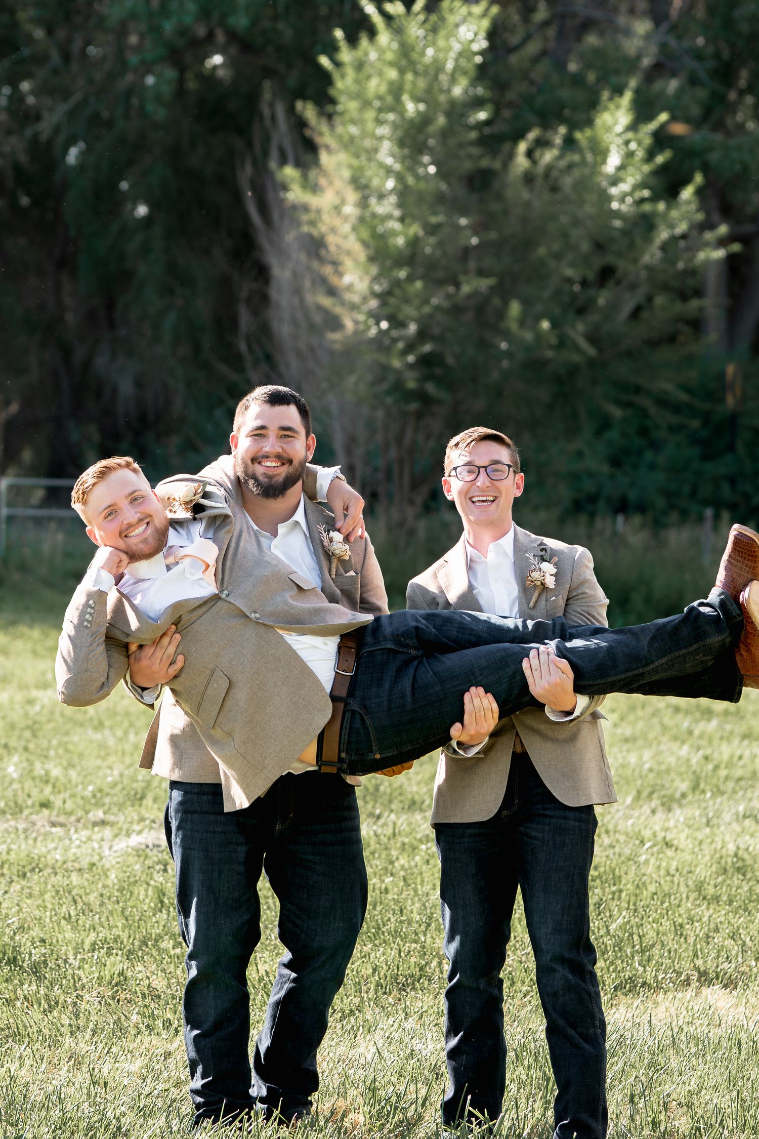 Groomsmen in gray suits playfully carrying and posing together on a sunny lawn.