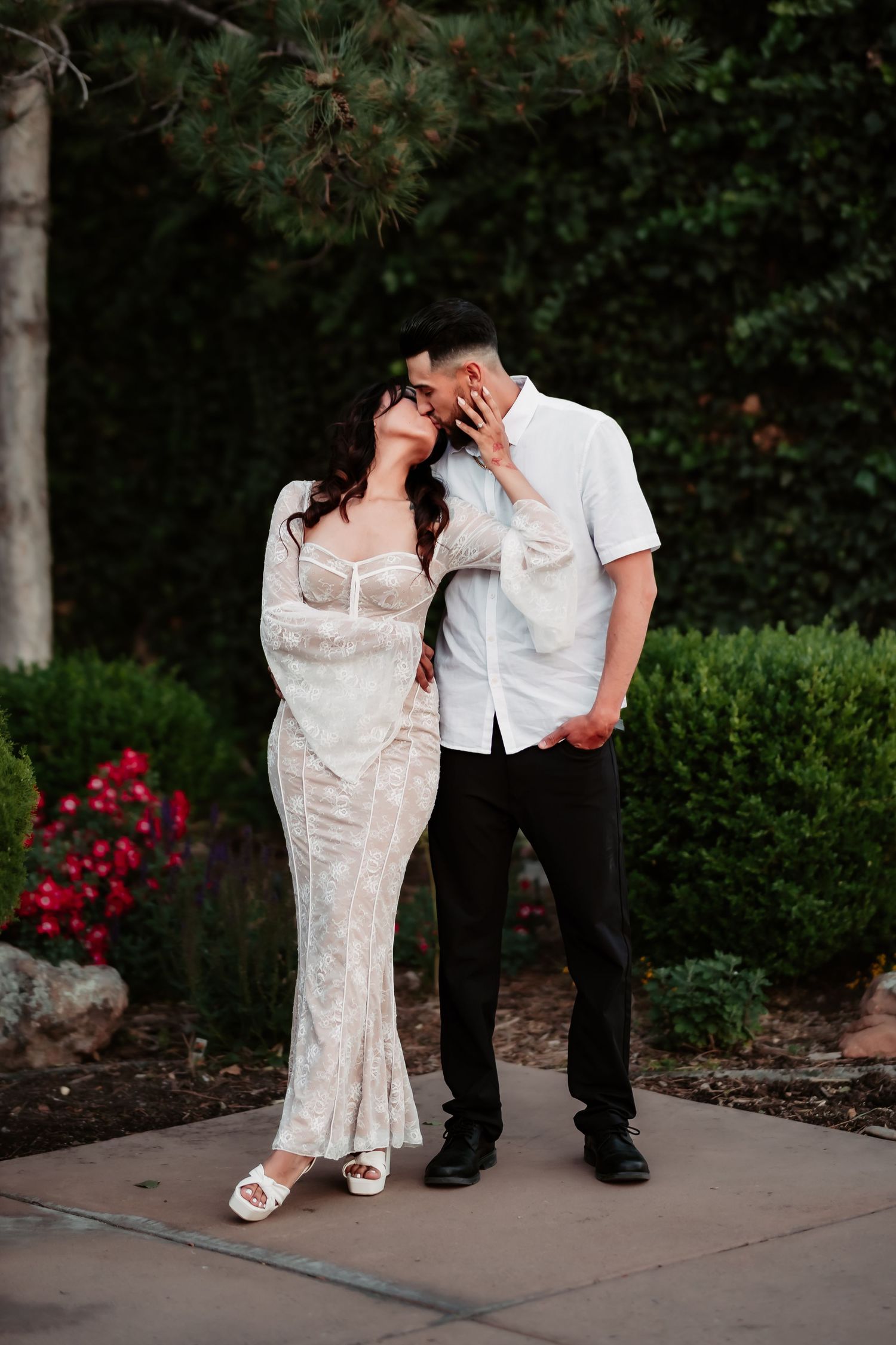 A romantic couple shares a close moment in a garden setting with red flowers and greenery.
