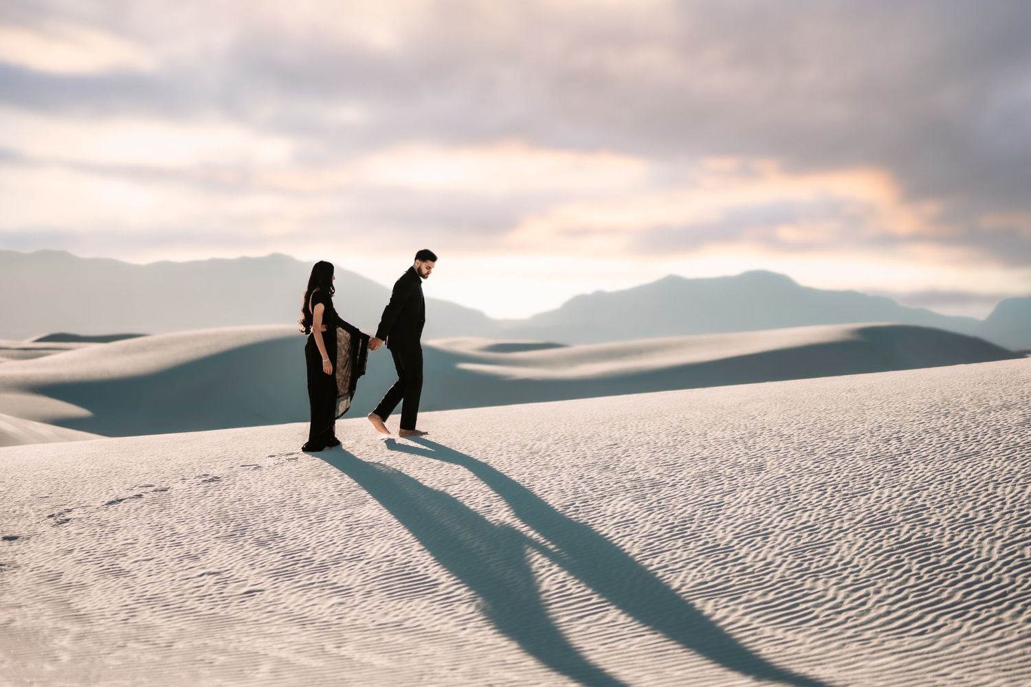 Two silhouettes walk along the white sand dunes at sunset casting long shadows against the pristine desert landscape.