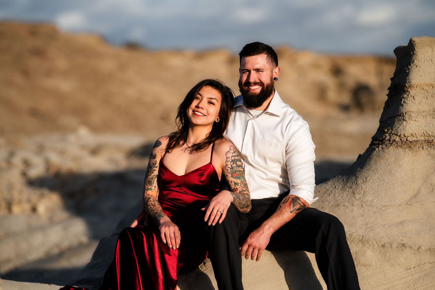 A couple in elegant attire poses intimately on desert rocks during golden hour.