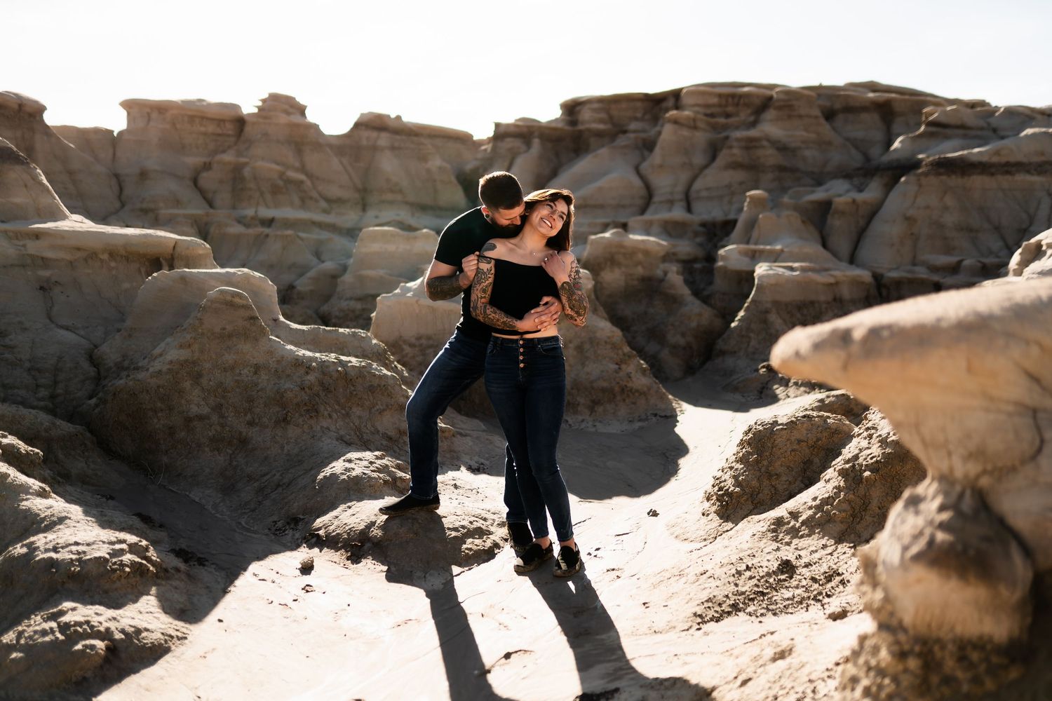 A couple embraces among unique rock formations in a desert landscape under natural sunlight.