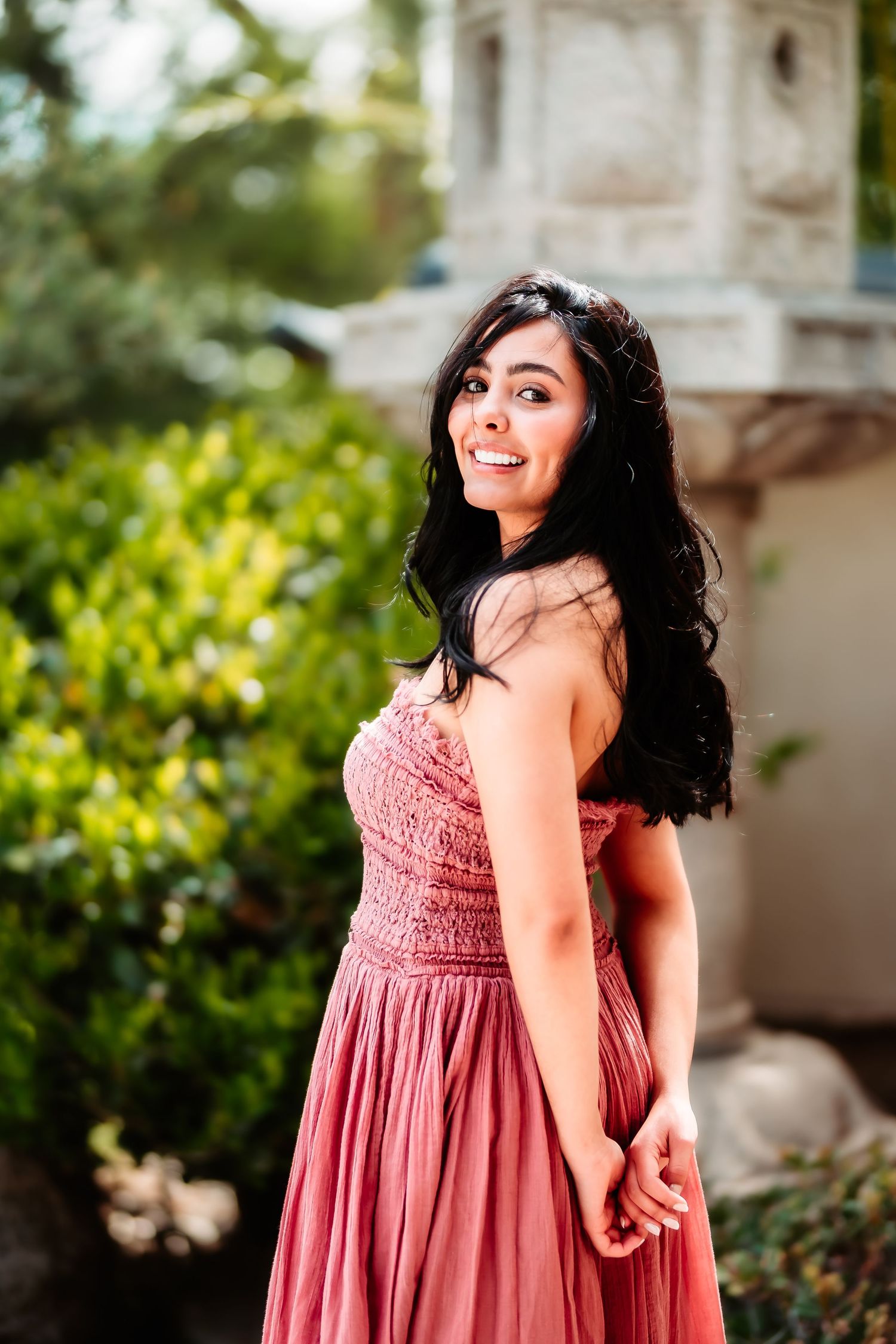 A woman in a flowing pink dress poses outdoors near a stone pillar and greenery.