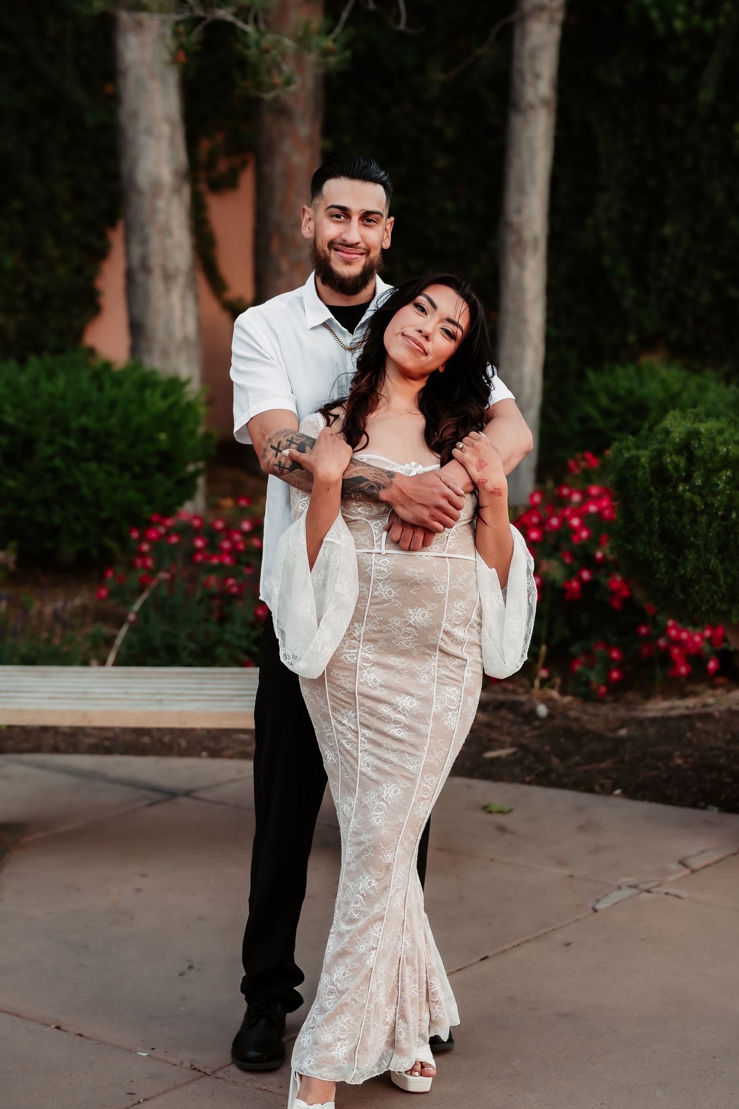 A romantic portrait of a couple embracing outdoors near blooming red flowers and trees at dusk.