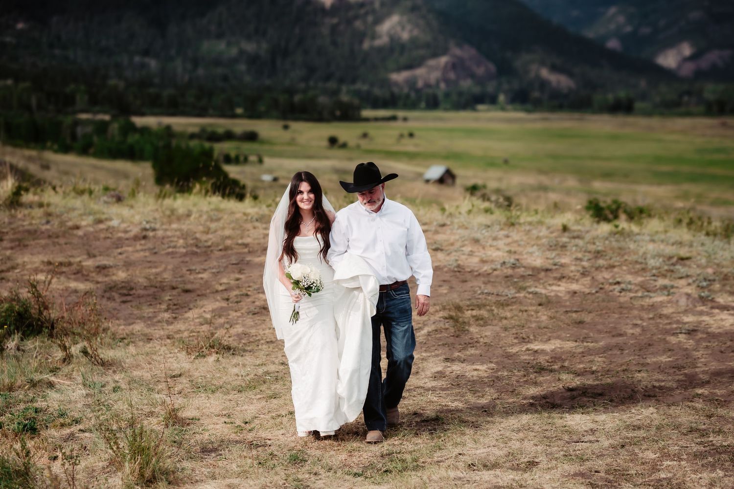 A couple walks together on a scenic mountain path with rolling hills and mountains in the distance.