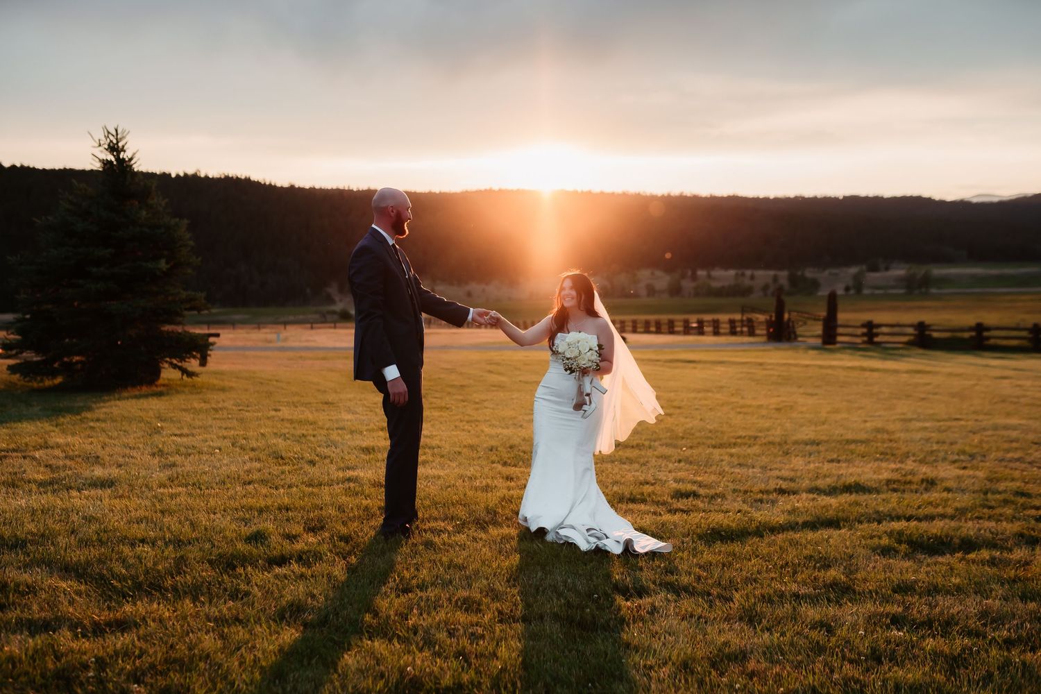 Wedding couple silhouetted against dramatic sunset in mountain meadow with evergreen trees.
