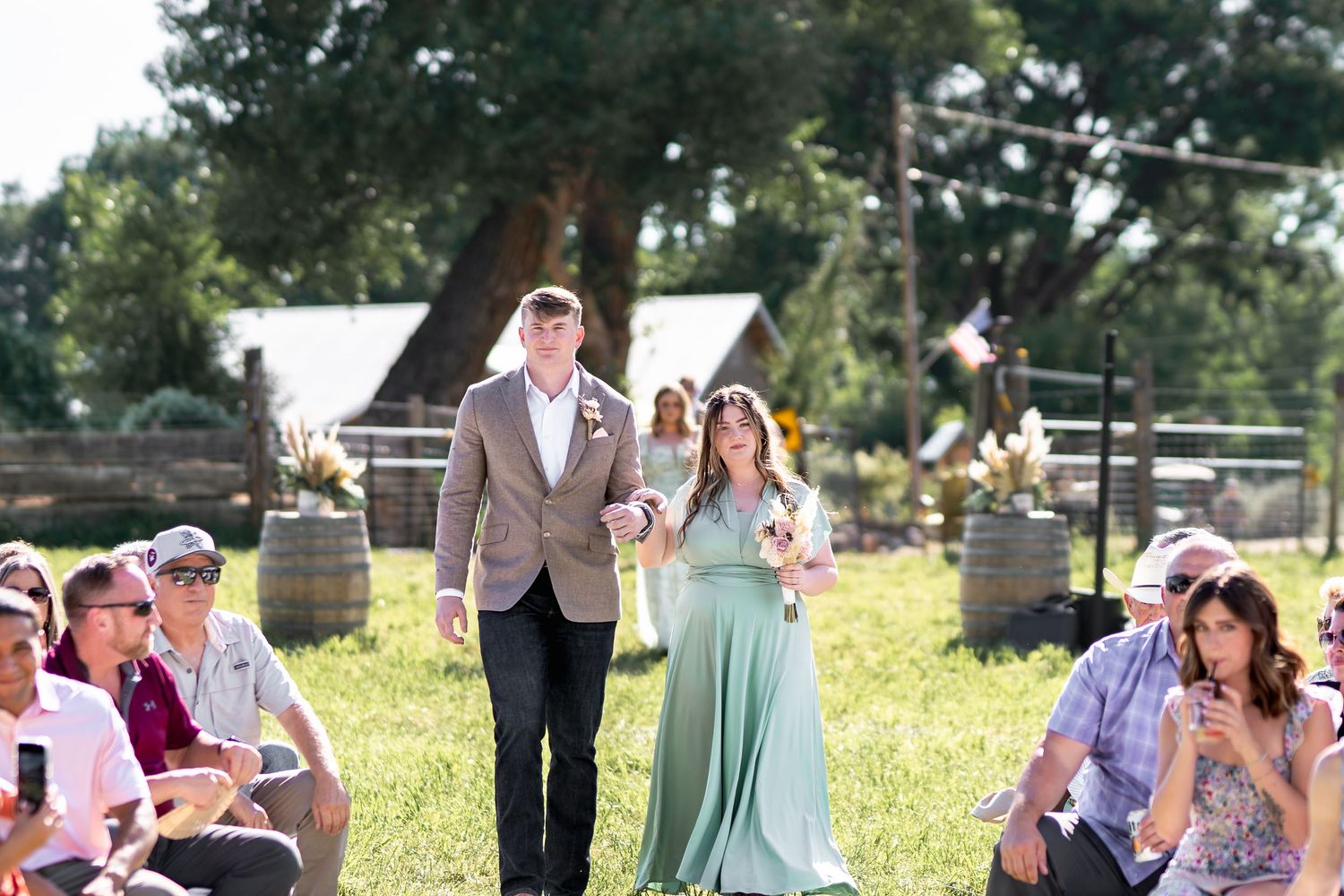 Wedding ceremony guests watch a procession on a bright sunny day with rustic barn in background.