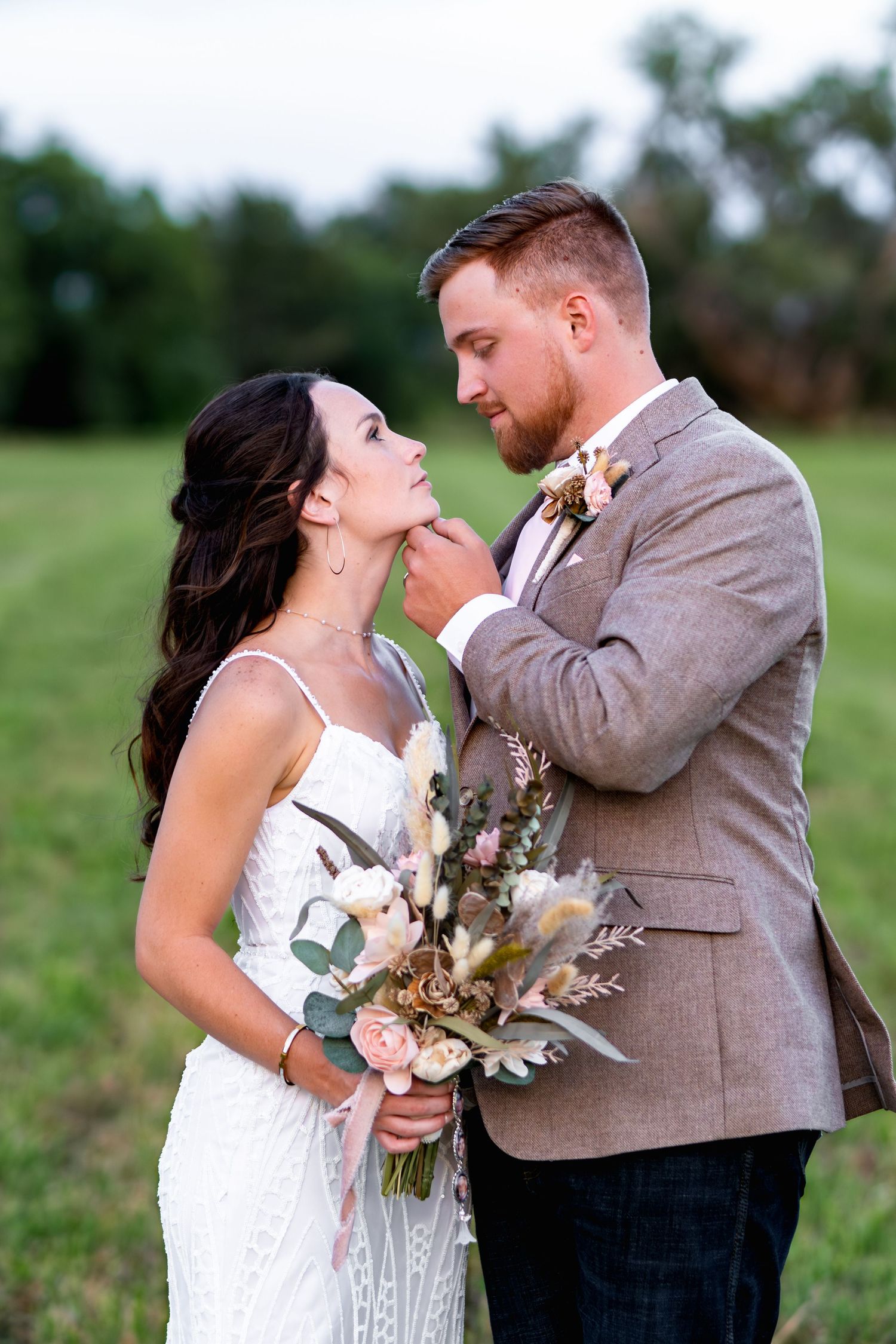 Romantic couple poses in outdoor field with elegant wedding attire and bouquet.