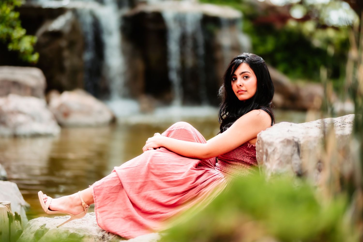 Woman in coral dress reclining on rocks near scenic waterfall.
