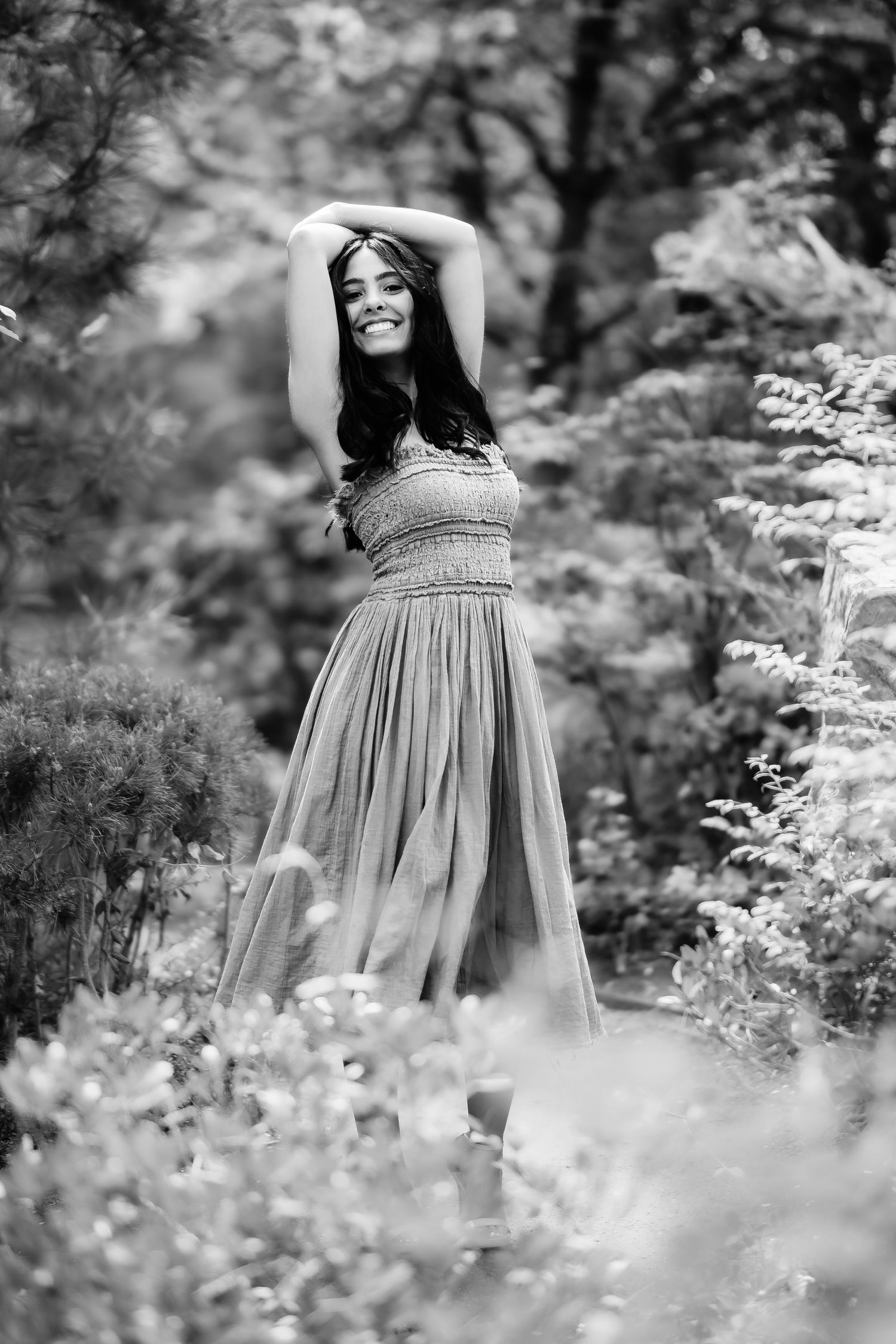 Black and white portrait of woman in flowing dress posing elegantly in garden setting.