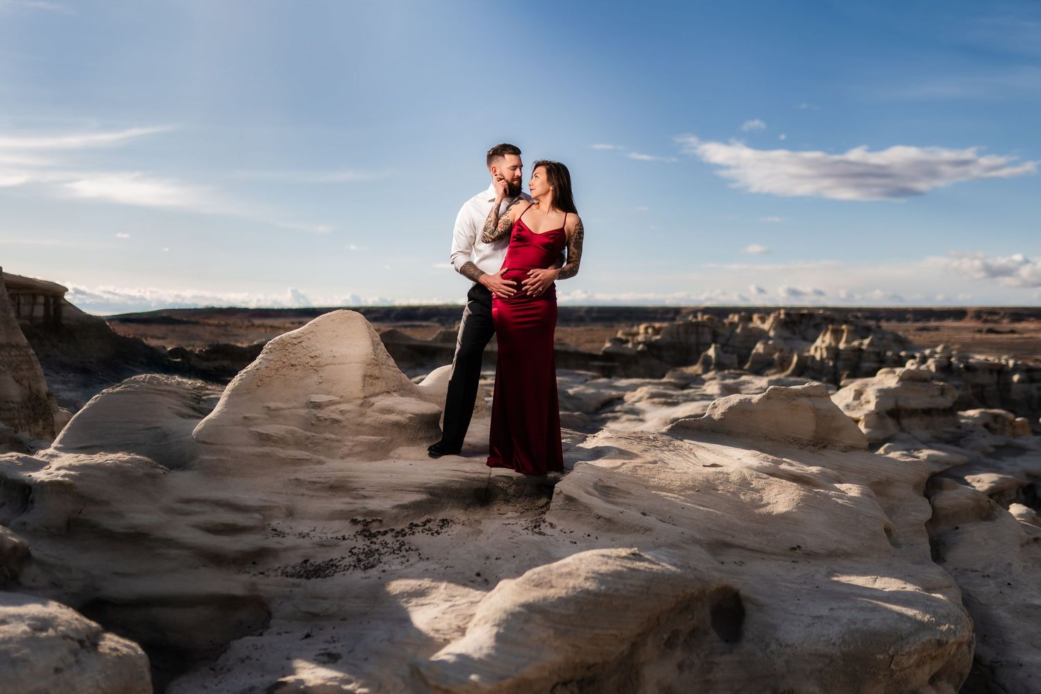 A romantic couple stands together on dramatic rocky terrain under a bright blue sky.