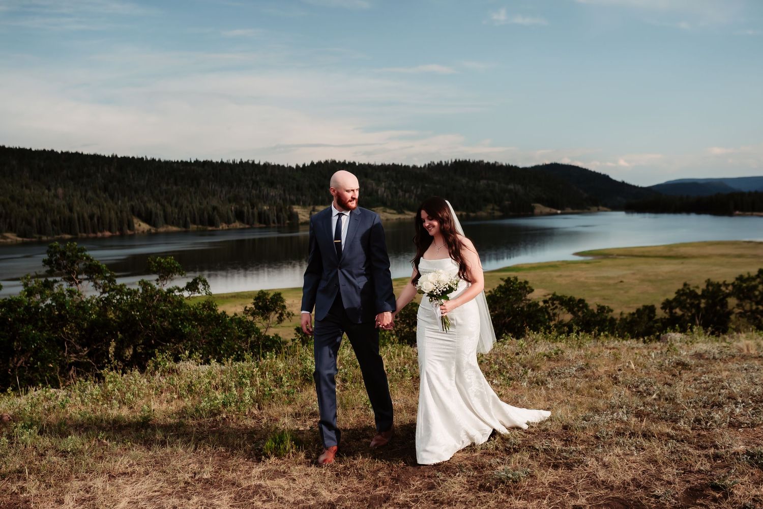 Newlyweds pose together on a hilltop overlooking a scenic lake and forest landscape.