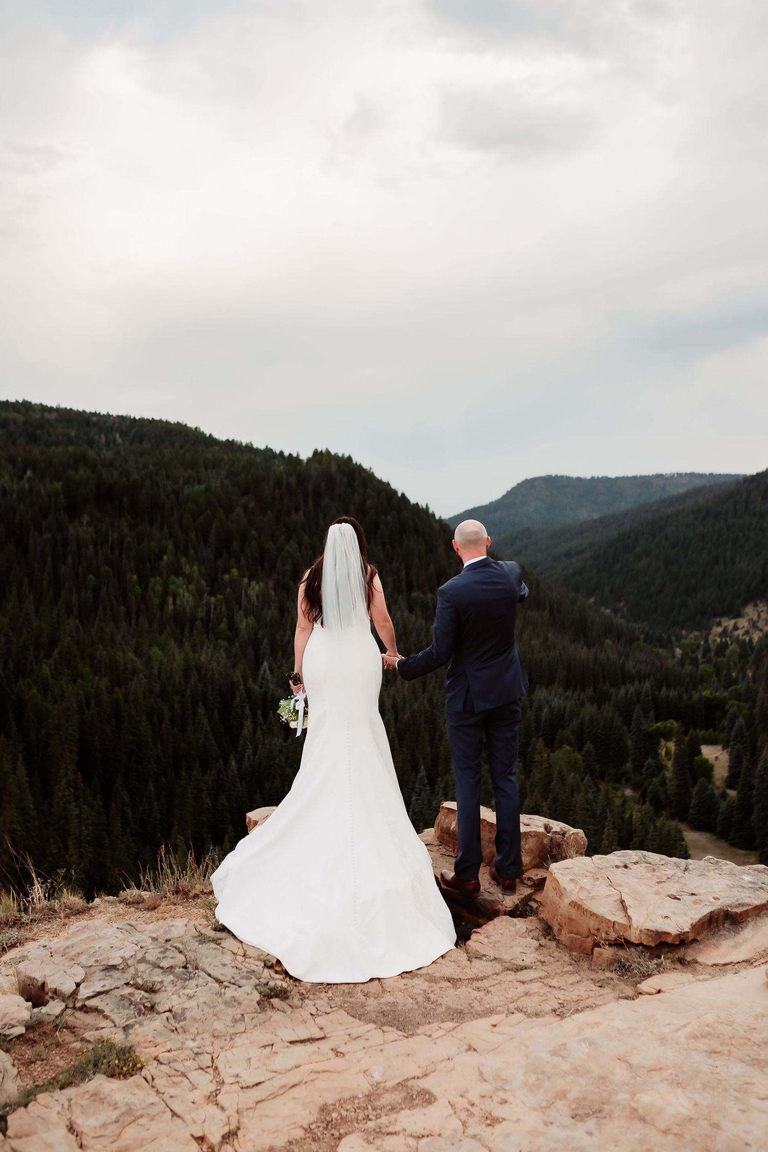 A couple in wedding attire stands together on a scenic mountain overlook with forested hills in the background.