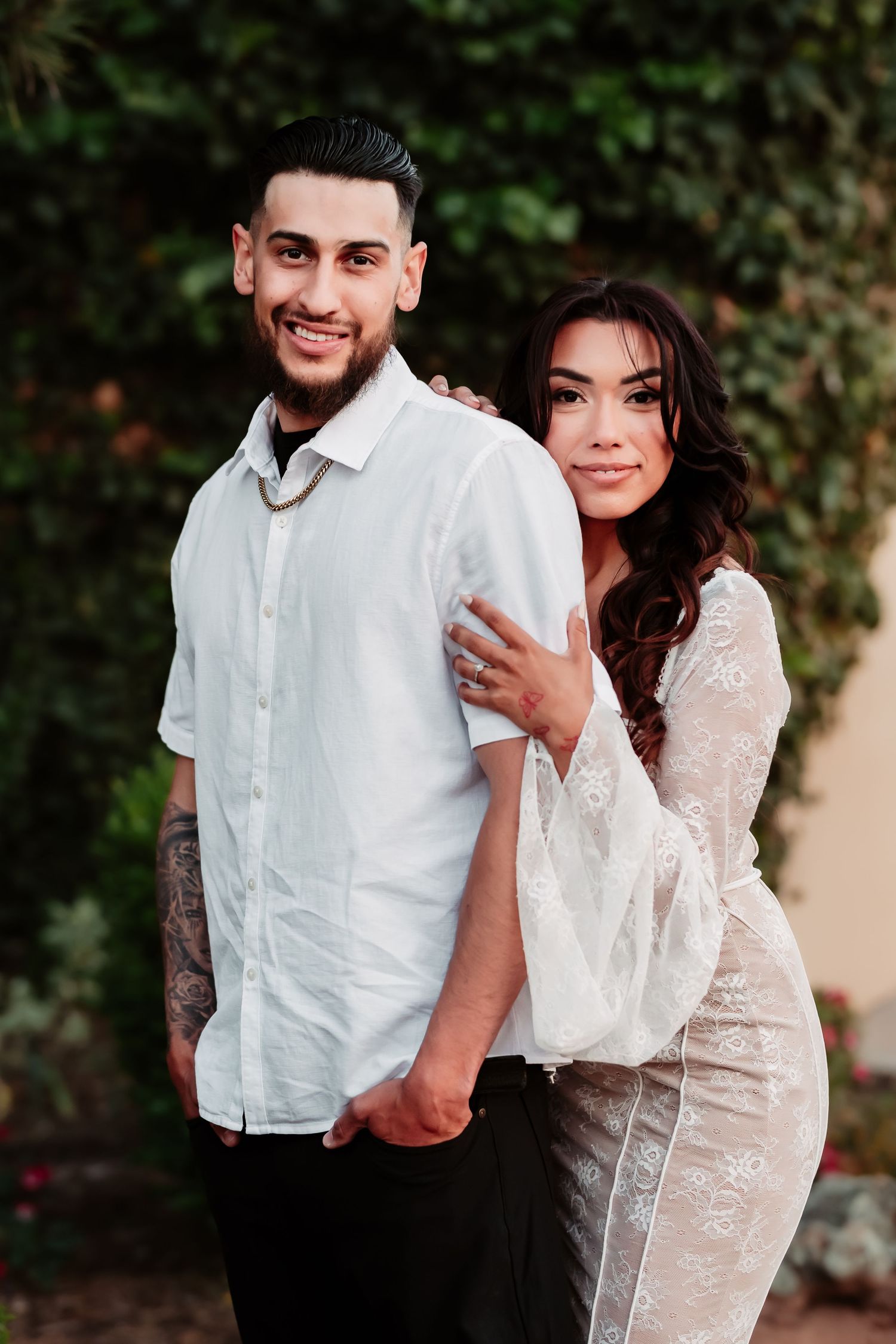 A romantic couple poses together in light colored outfits against a garden backdrop.