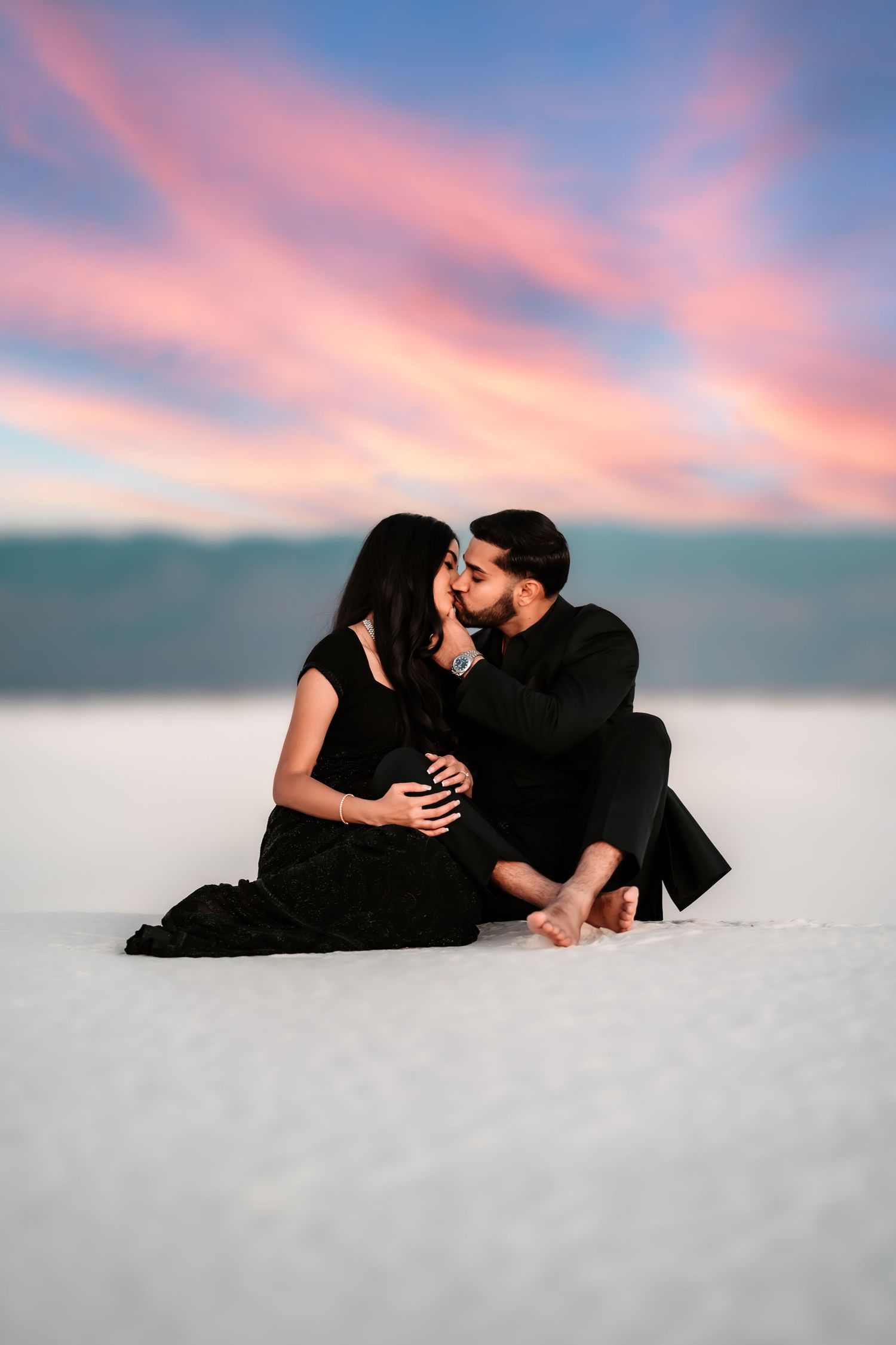 A couple in black outfits shares an intimate moment on white sand under a pink and purple sunset sky.