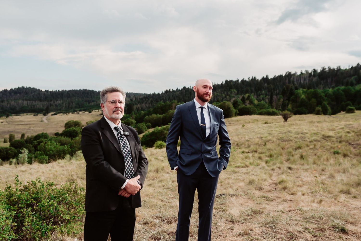 Wedding party members in formal attire gathered outdoors with mountain scenery in background.