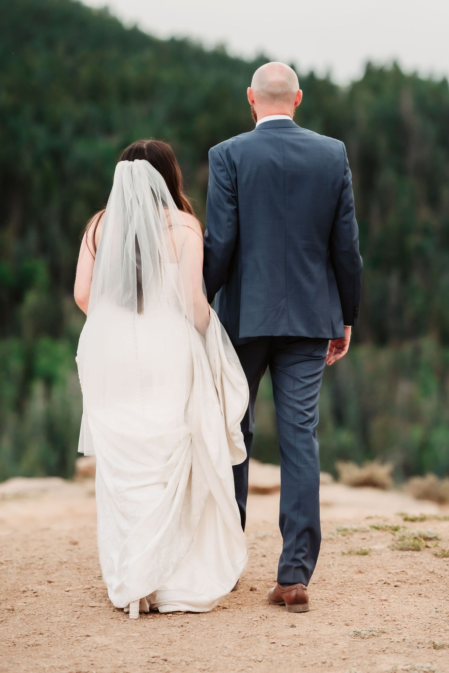 Newlywed couple walking together on a sandy path with dense forest in the background.