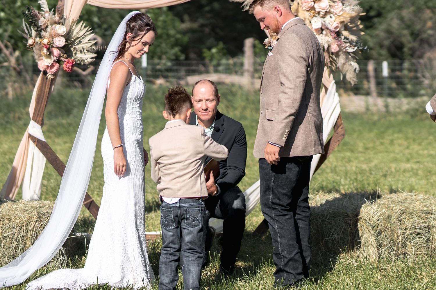 An intimate outdoor wedding ceremony under a floral arch with a young ring bearer joining the couple.